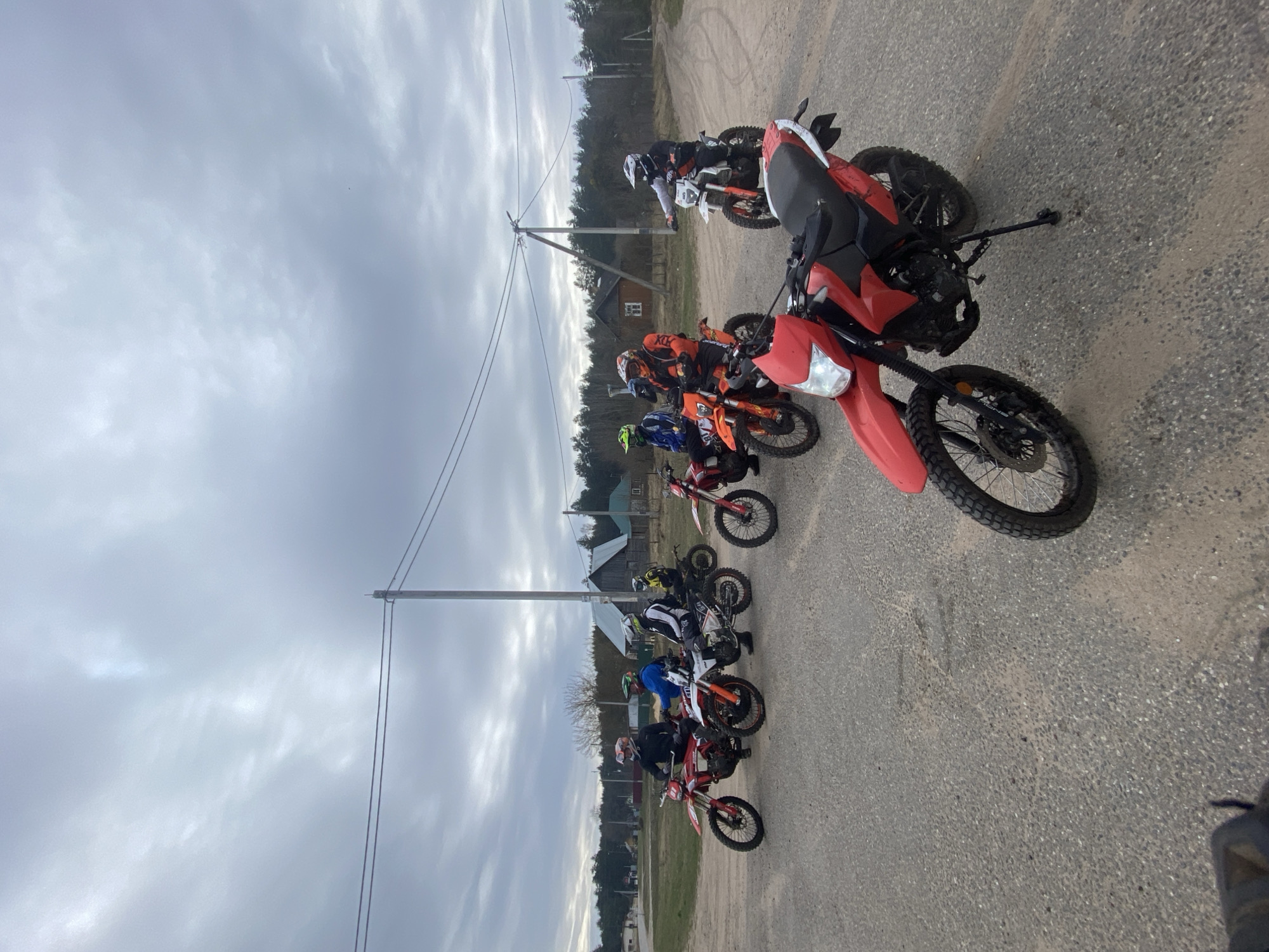 The image shows a group of approximately 8-10 people on dirt bikes parked on a paved road in Гожскі сельскі Савет, Belarus. The bikes are a mix of red, orange, and white models. The riders are wearing helmets and riding gear. In the background, there are power lines and houses. The sky is overcast.