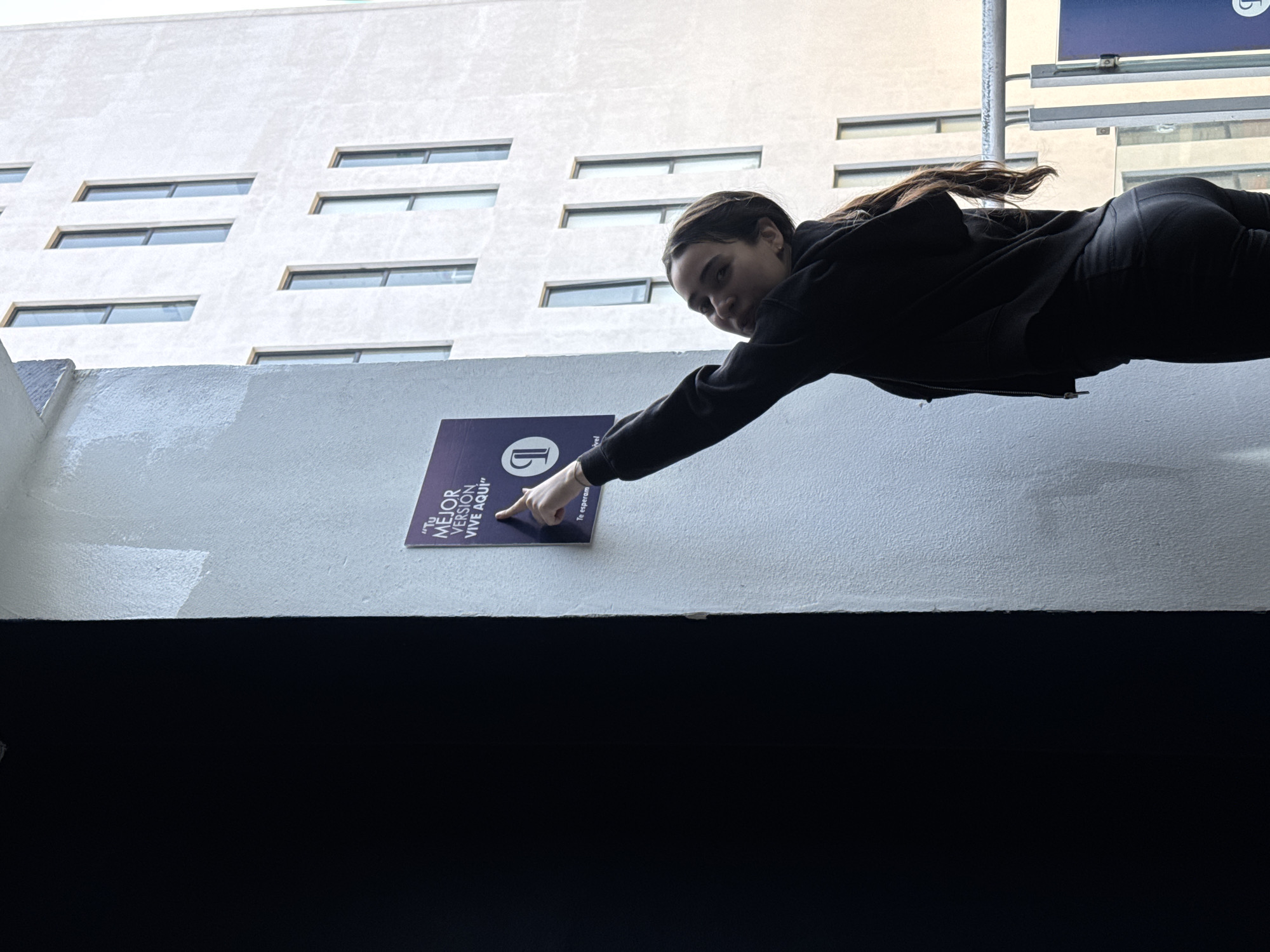 A young woman is captured in a striking pose, appearing to float horizontally in the air while reaching out to touch a sign on a wall. She is positioned on the right side of the frame, dressed in a dark (likely black) long-sleeved top and matching pants, with her dark hair pulled back. Her face is visible as she looks towards the camera with a subtle smile.
Her right hand extends to a rectangular sign mounted on a textured white wall. The sign is purple or dark blue with white text. Visible text on the sign reads: "TU MEJOR VERSIÓN VIVE AQUÍ" (Your best version lives here) and below that, "Te esperan" (They await you). The sign also features a white stylized logo, possibly a 'G' or similar shape.
Below the white wall is a dark, undefined horizontal surface. Above the wall, a tall, light-colored building with a textured facade dominates the upper half of the image. This building features multiple horizontal rectangular windows arranged in an irregular pattern across its surface. The photograph is taken from a low angle, emphasizing the upward perspective and contributing to the illusion of the woman's horizontal position. The bright, even lighting suggests it is daytime with clear weather. This scene takes place in Puebla, Mexico.