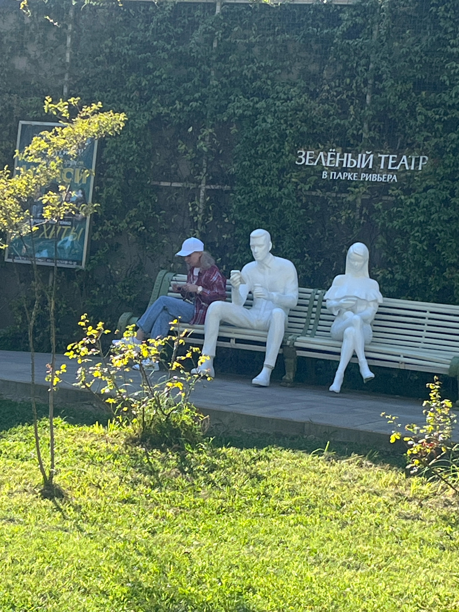 The image shows the Green Theatre in Riviera Park, Sochi, Russia. A woman wearing a white baseball cap, a red and black plaid shirt, and blue jeans is sitting on a bench, looking at her phone. To her right are two white statues of people sitting on the same bench. The statue on the left is of a man holding a phone, and the statue on the right is of a woman holding a book. The bench is positioned in front of a wall covered in ivy. In the foreground, there is a grassy area with some bushes. A sign with text in Russian is partially visible on the left side of the image, behind a tree.