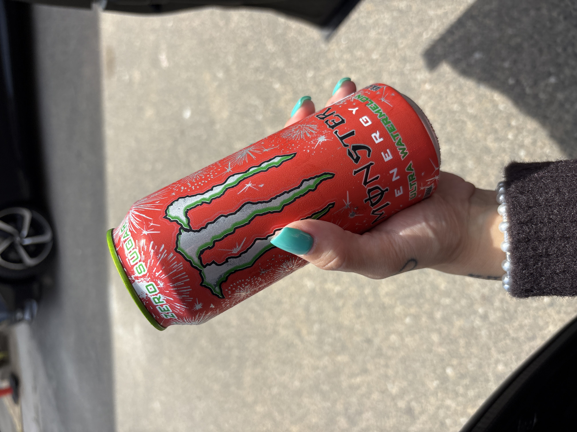 A person with turquoise manicured nails is holding a red and silver Monster Ultra Watermelon energy drink can. The can has the "ZERO SUGAR" label on the bottom. The person is wearing a dark, possibly knitted, sweater with a pearl bracelet. The background shows a concrete surface with shadows and a car wheel. This image was likely taken outside near a vehicle. There is no information in the image that directly points to Huntington Beach, United States of America. The inclusion of Huntington Beach would have to be extrapolated from other information.