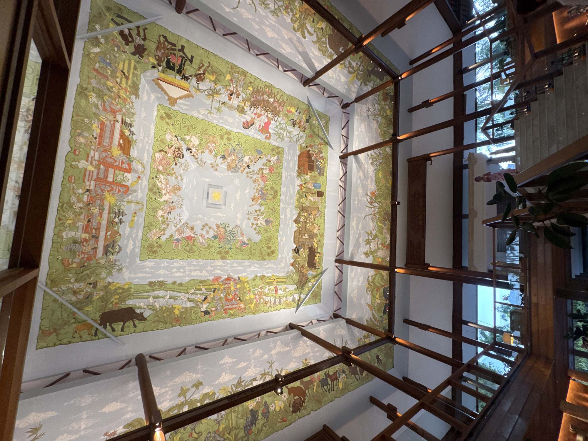An eye-level, wide indoor shot looks straight up to a colorful painted ceiling and an overhead structure of long, dark brown beams in the Apurva Kempinski Hotel, Kuta Selatan, Bali, Indonesia. The painting is a pale blue with green borders containing painted figures, animals, and architectural images. The center of the painting has a yellow star in a square. The beams intersect, and have small lights on them. Through a window to the side is a set of stairs. There are green plants in the right side of the photo.