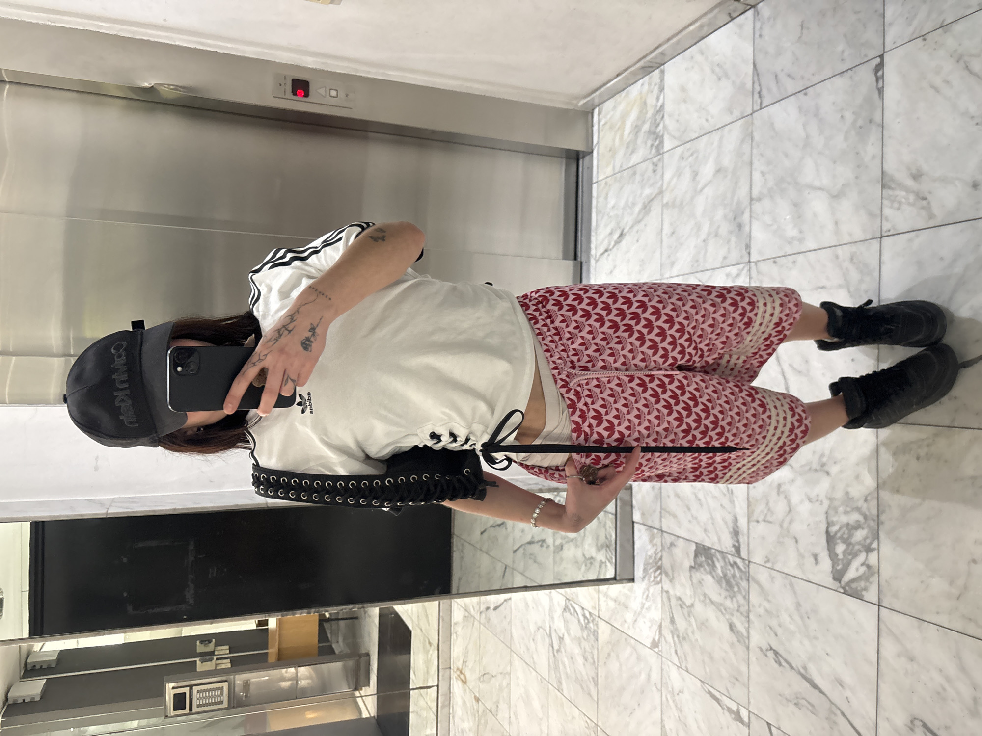 A person is taking a mirror selfie in front of an elevator. The person is wearing a white Adidas t-shirt with black stripes on the sleeves, pink patterned pants, and black boots. They have a black baseball cap on their head and are holding a black smartphone in their hand. A black bag is slung over their shoulder. The elevator is stainless steel. The floor is made of white marble tiles. Part of a kitchen is visible in the mirror's reflection. The city location, Buenos Aires, Argentina, cannot be determined from the image.