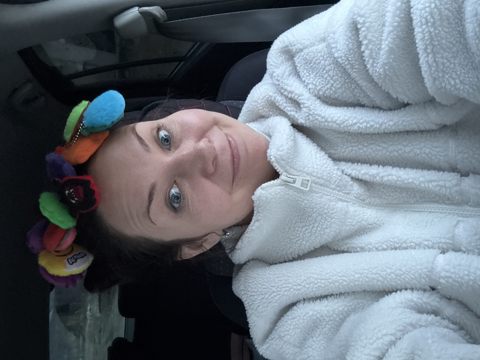 The image is a close-up, sideways selfie of a woman positioned inside what appears to be a car. She is the central subject, looking directly at the camera with wide blue eyes and a gentle, slight smile. Her expression conveys contentment or amusement.
The woman has dark hair and is wearing a thick, white, textured fuzzy jacket, resembling sherpa or fleece, with a white zipper visible down the front. Hints of dark clothing are visible underneath the jacket. On the left side of her head, she sports a vibrant, colorful accessory, possibly a headband or hair clip, adorned with multiple small, fluffy, round charms or pom-poms. These charms are in various hues, including blue, green, orange, yellow, purple, and red. One yellow charm displays a small, blurry, and mostly illegible text or logo, while a red charm features a simplified cartoon-like face.
The background elements, including a car seatbelt, the dark interior headliner, and a window frame, indicate the setting is inside a vehicle. The lighting is dim, suggesting it could be evening, nighttime, or a heavily overcast day. While specific weather conditions are not visible, the cozy jacket implies cooler temperatures. The user indicates the location is Chaikovsky, Russia.