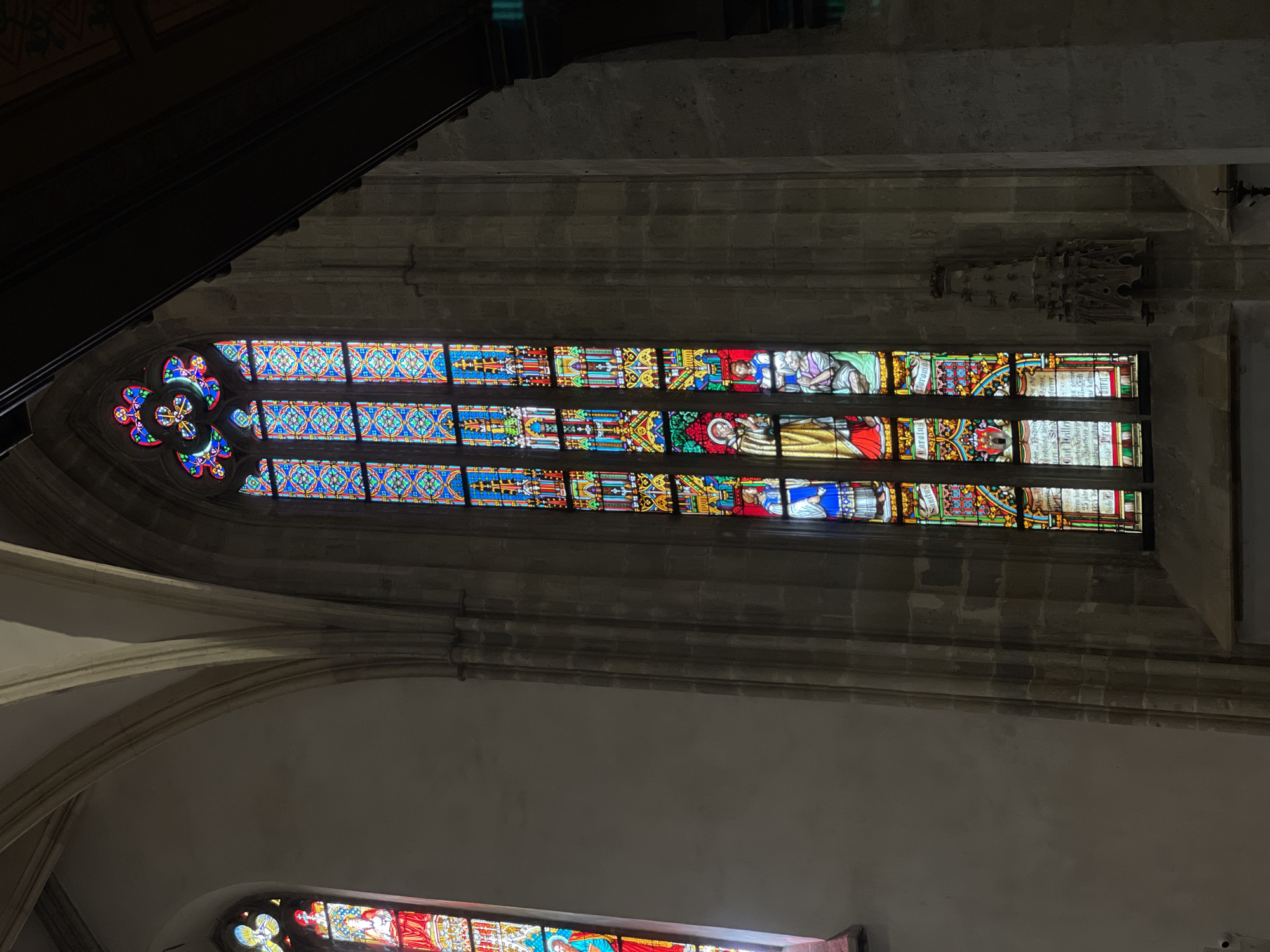 This image captures a close-up view of a tall, narrow stained-glass window in a dimly lit interior, likely a church or cathedral, given the architectural style and the presence of stained glass. The window is divided into multiple horizontal panes, each filled with intricate designs and scenes rendered in vibrant colors, including blues, reds, yellows, and greens. The leftmost section of the window features a circular rose window design with four pointed lobes, each filled with detailed patterns. The horizontal panes above and below the rose window depict various figures, possibly biblical or historical characters, engaged in different activities. Some figures are depicted with halos or elaborate robes, suggesting religious or noble status. The stained glass is set within a stone frame, characteristic of Gothic or Romanesque architecture. Above the main window, dark, angled beams suggest a vaulted ceiling or roof structure. The overall lighting is low, with the stained glass being the primary source of illumination and color. The scene evokes a sense of historical and artistic grandeur, common in European religious institutions. The image is taken from below, looking upwards towards the window. No specific time of day or weather conditions are discernible.