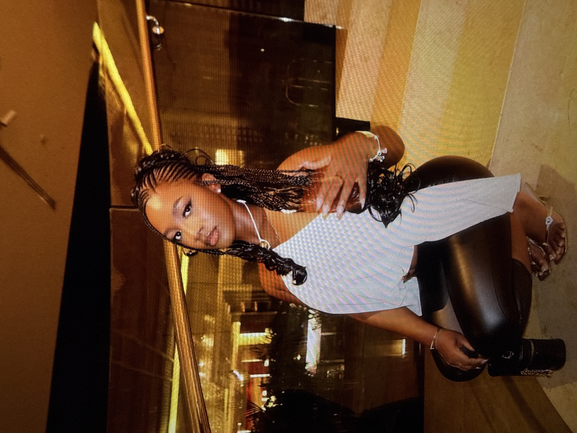 A woman with dark skin and braided hair is posing for a photograph, crouched down in a well-lit indoor space, possibly a hotel lobby or restaurant, with warm-toned lighting and reflective surfaces. She is wearing a light-colored halter-neck top and black leather pants. Her right hand is raised and holding a champagne flute, while her left hand rests on her knee. A small black handbag is placed on the floor beside her left leg. The background features a blurred view of interior lighting and possibly outdoor scenery seen through a window or glass partition. The overall impression is one of an elegant evening setting.