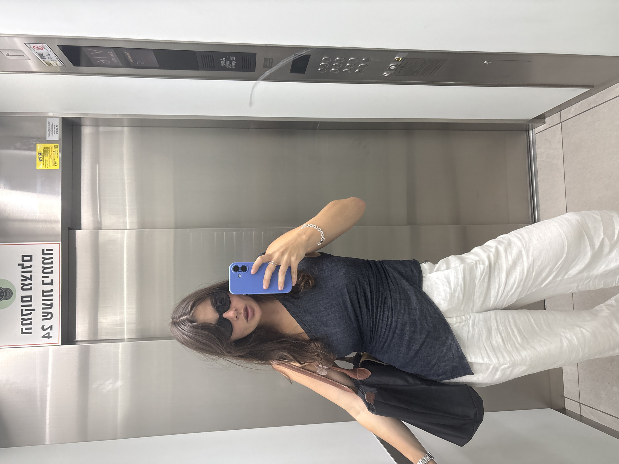 In an elevator lobby, a young woman with long brown hair, wearing sunglasses, a dark one-shoulder top, and white wide-leg pants, takes a selfie with a blue phone. She is carrying a black tote bag on her left shoulder. The elevator doors are made of stainless steel, and there is a control panel on the right with buttons. A sign with Hebrew text is visible above the elevator doors. The floor is tiled.