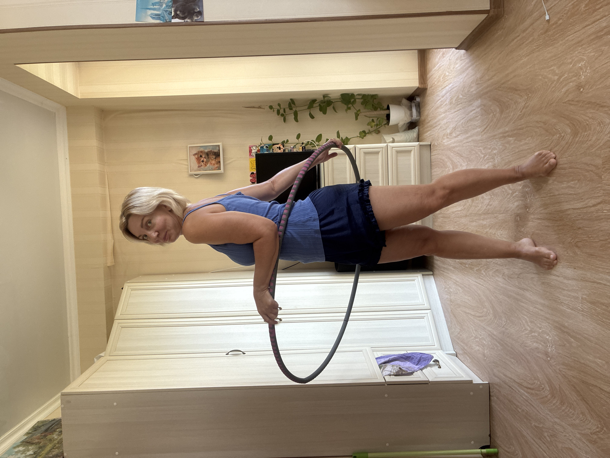 An adult female with blonde hair, wearing a blue tank top and dark blue shorts, stands in a room and holds a hula hoop. She is looking over her left shoulder towards the camera with a slight smile. The room appears to be a bedroom or living area, with a large wardrobe on the left, a dresser and television in the background, and a large mirror on the right. There are plants and decorative items on shelves and on the floor. The flooring is light-colored wood laminate. The overall impression is that of a casual indoor setting. This scene is not specific to Sochi, Russia, and lacks any discernible landmarks or indicators of that location.