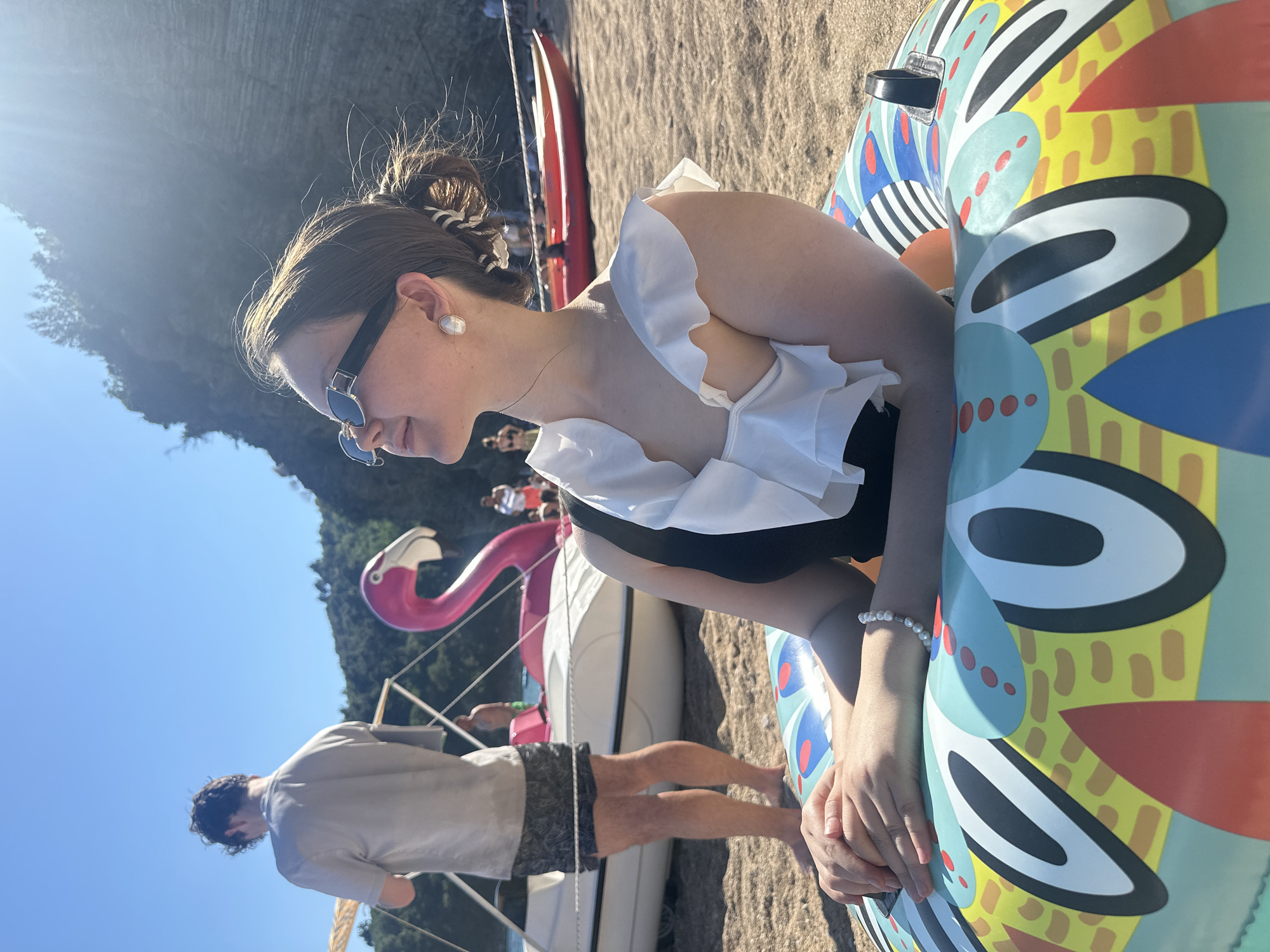 A daytime shot on a beach in Budva, Montenegro, shows a young woman in sunglasses and a white ruffled top with a black bottom sitting in an inflatable raft with a colorful design. Her hair is in a bun, and she wears pearl earrings and a bracelet. To her left and behind are a pink flamingo decoration, a white boat, and a man in shorts and a grey shirt. People are standing in the background. The location is sandy with a hill or mountain in the distance. The sky is blue, and the sun is shining.