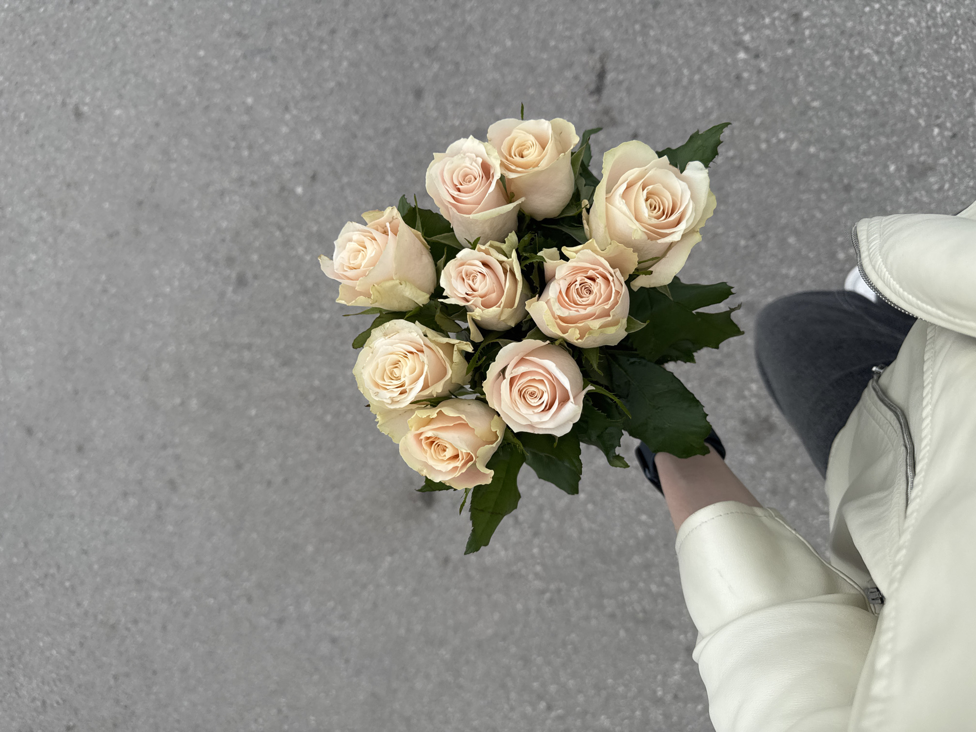 Reportedly located in Perm, Russia, the image captures an overhead view of a person holding a bouquet of roses over a paved outdoor surface. The ground is a uniform grey, granular texture consistent with asphalt or concrete, displaying subtle variations in shade and small, scattered specks. A person's left arm, wearing a light cream or off-white jacket with a visible zippered detail on the torso, extends from the lower left, holding the bouquet. Part of a dark grey leg, presumably covered by trousers or jeans, is visible on the lower right, with a glimpse of a white shoe. The bouquet consists of approximately nine pale peach and cream-colored roses, each with fully formed, spiraled petals. Dark green leaves are interspersed among the blooms, providing a contrasting backdrop.