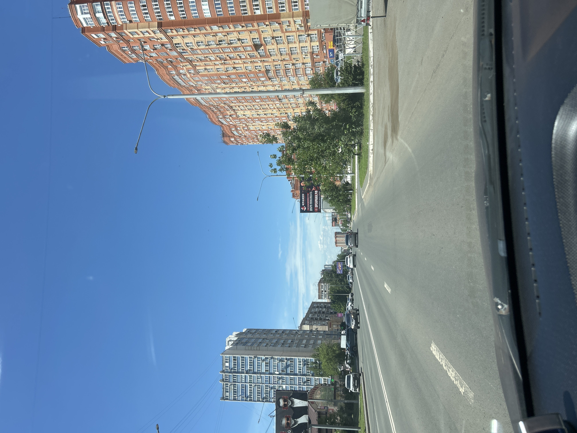 The image depicts a sunny day on a multi-lane road in Orenburg, Russia. On the left side of the road stands a tall, light blue apartment building with numerous windows and balconies. A billboard featuring two men in sunglasses is positioned in front of this building. Further down the road, smaller buildings and trees are visible. On the right side of the road, a large, modern apartment building with orange and yellow brickwork dominates the view, characterized by its curved facade and many windows. Streetlights line the road. Several cars are driving on the road, including a white sedan, a dark SUV, and a white sedan. A white truck with a trailer is parked on the right side of the road. The sky is clear blue with a few wispy clouds. The foreground shows the dashboard of a vehicle, indicating the image was taken from inside a car.