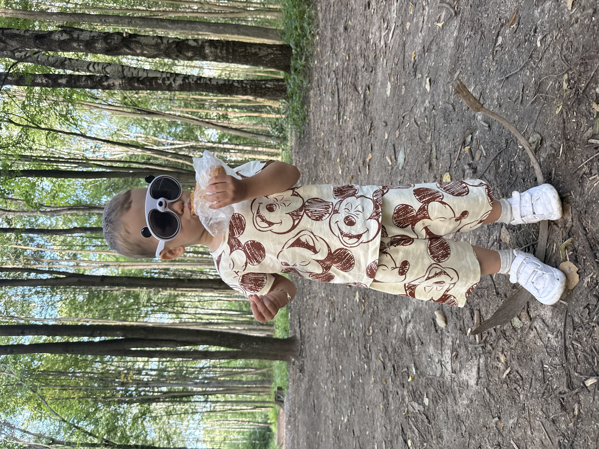A young child, wearing sunglasses with panda ears and a light-colored t-shirt and shorts with a Mickey Mouse pattern, stands in a forest in Rusanovo, Russia. The child is holding a pastry wrapped in plastic and is in the process of eating it. The ground is covered with dirt, leaves, and small branches. Tall trees with green foliage surround the child, and the overall lighting suggests it is daytime.