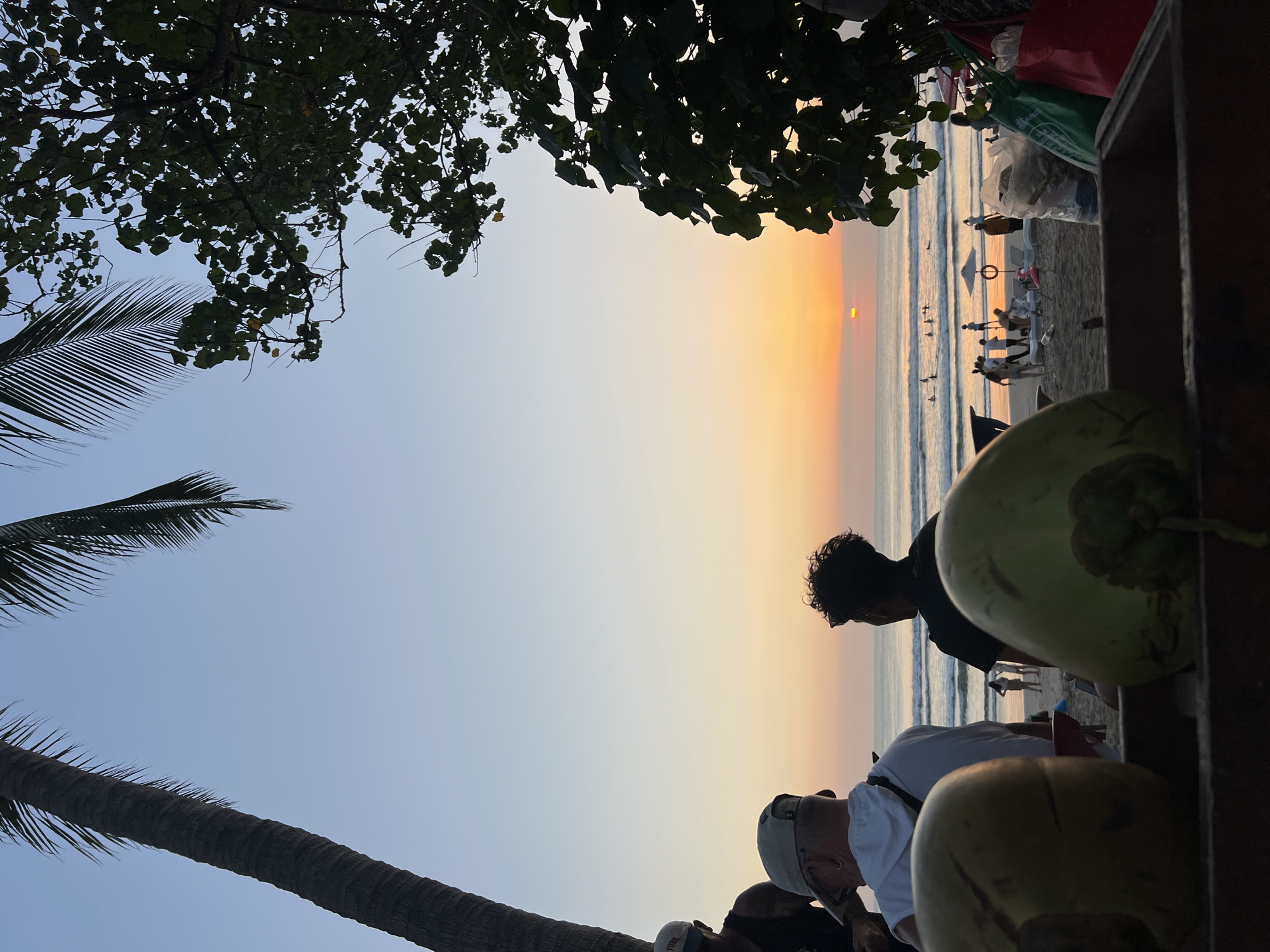 This is a low-angle, close-up shot of coconuts in the foreground, positioned in front of a beach scene in Kuta, Indonesia during sunset. Two large, green coconuts are visible, with their textured brown husks at the top. Behind the coconuts, a group of people are gathered on the sand, facing the ocean. Several individuals are walking, standing, or sitting. Further back, the ocean is calm with gentle waves and several surfers are visible in the water. The sun, a bright orange disc, is setting on the horizon, casting a warm glow across the sky and water. Palm trees and leafy branches frame the scene from the left and right, respectively. The overall atmosphere is one of a relaxed beach setting at dusk.