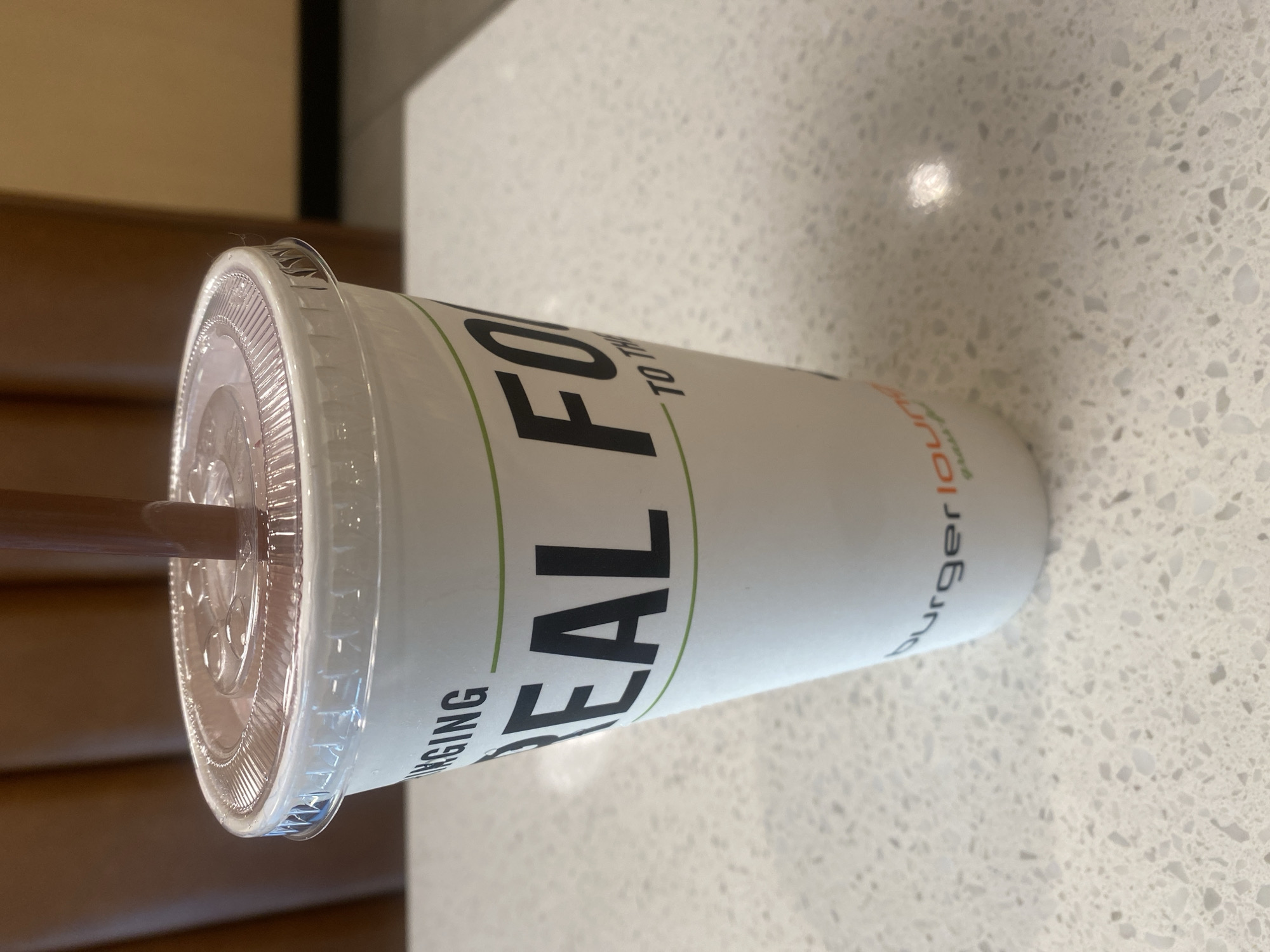 A close-up shot shows a white paper cup with a clear plastic lid and a brown straw. The cup has black text that reads "EAL FO" in large font, with smaller text above and below. Above, "NGING" is visible, and below, "TO TH" is visible. The brand name "burger lounge" appears in a curved font near the bottom of the cup, accompanied by green lettering. The cup is positioned on a speckled white countertop. The background consists of blurred brown and tan surfaces, suggesting an indoor environment. This image was taken at Burger Lounge in Manhattan Beach, United States of America.
