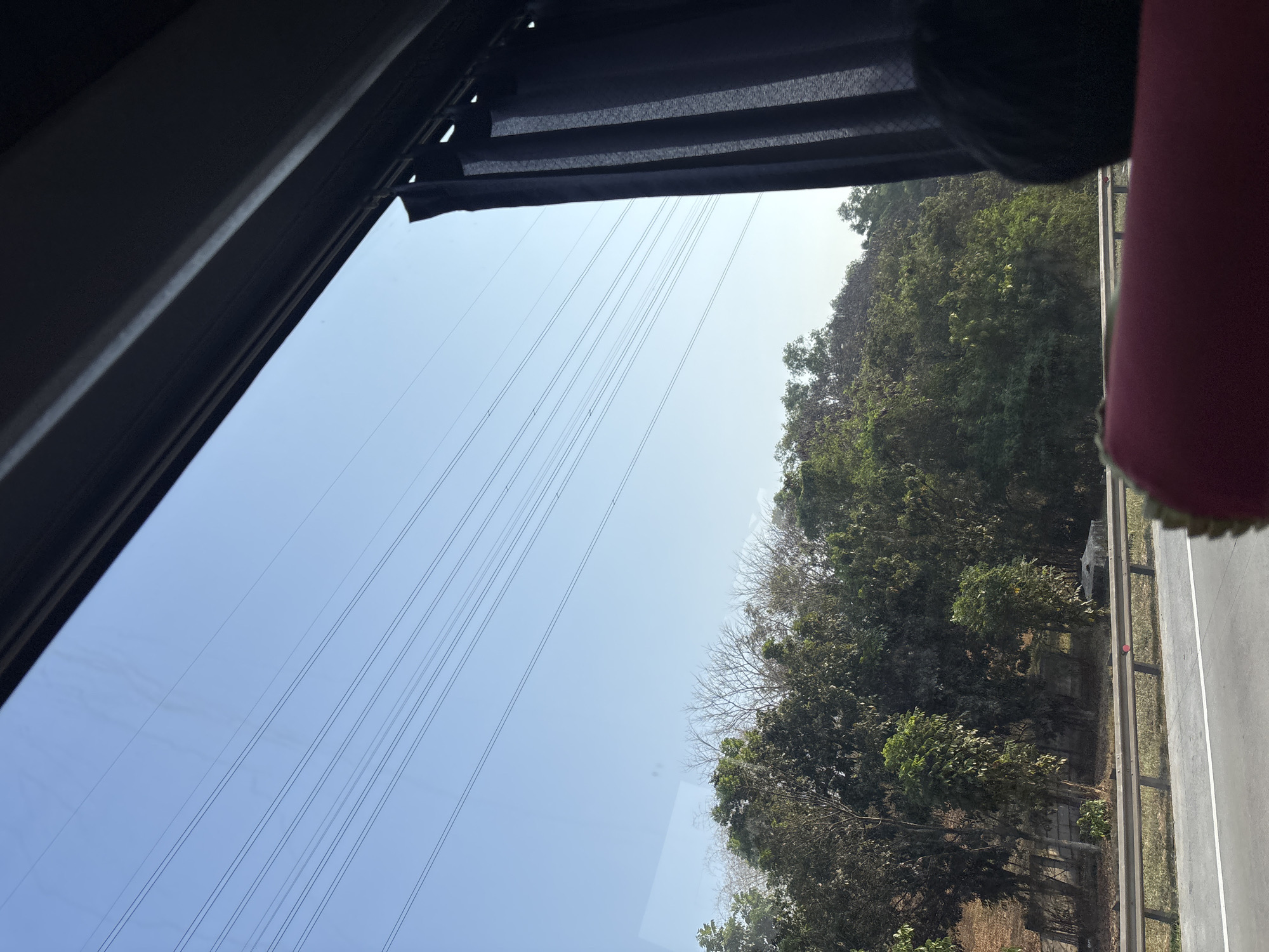 This image depicts a view from inside a vehicle, looking out through a window. The dominant elements are the clear blue sky, intersected by multiple parallel power lines. Below the sky is a dense line of trees and foliage, suggesting a natural or semi-rural environment. In the foreground, a dark curtain is visible on the right side, partially obscuring the view. A portion of a red seat or cushion is also in the very bottom foreground. A guardrail separates the road from the embankment where the trees grow.
While the image shows a natural landscape with infrastructure, there is no specific visual information within the image that allows for the identification of the city of Karawang, Indonesia, or any specific location within it. The scene is generic to many areas with power lines, trees, and a road.
