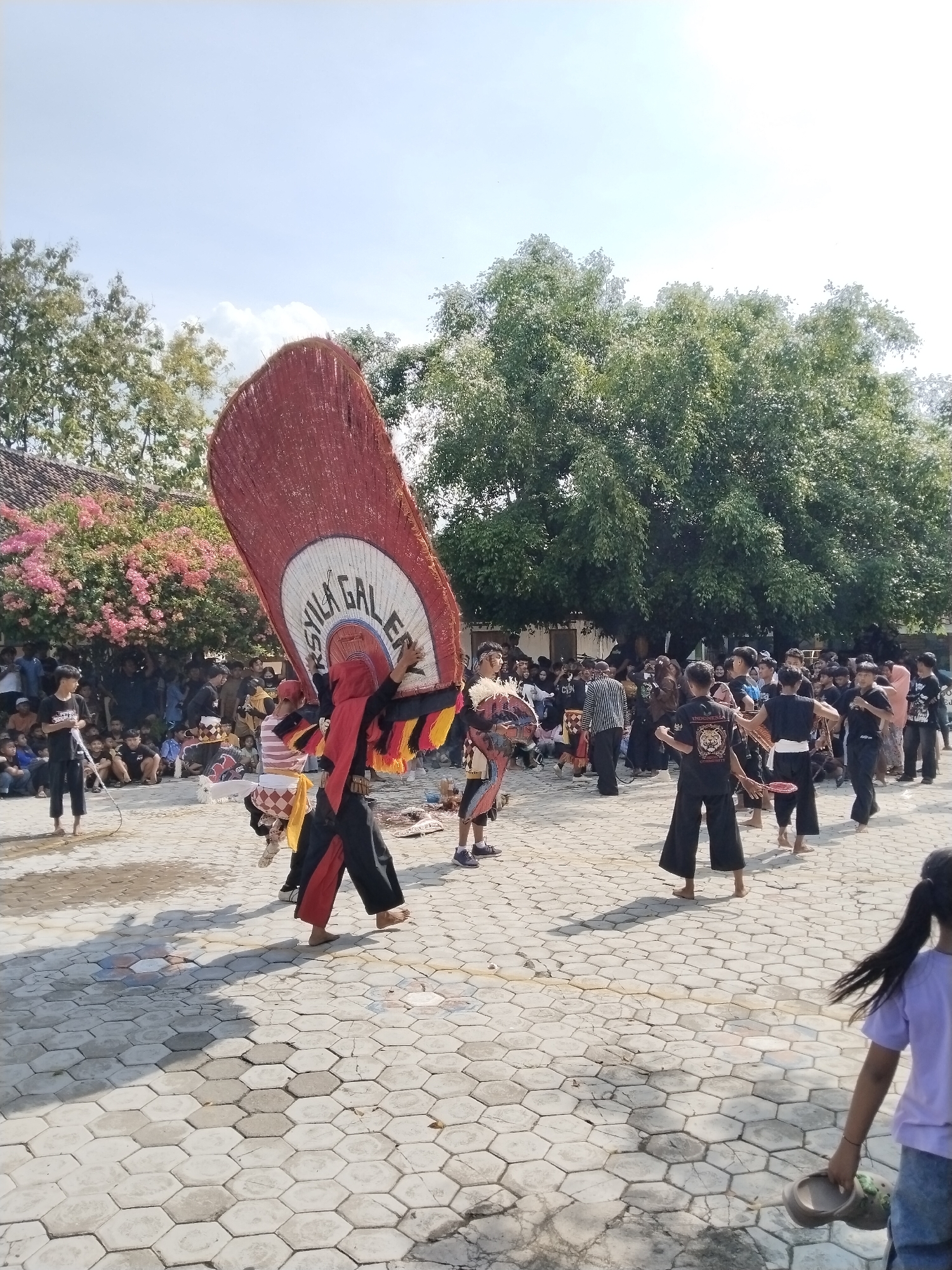 The image captures a cultural performance in Karangmalang, Indonesia. A performer wearing traditional attire and a large, decorated prop with the words "ARSYILA GALLERY" is the central figure. Other performers, dressed in black pants and some shirtless, are participating in the event. Many people are observing the performance, some seated and others standing in the background. The setting appears to be an outdoor paved area, with trees and foliage in the background. The sky is clear and blue.