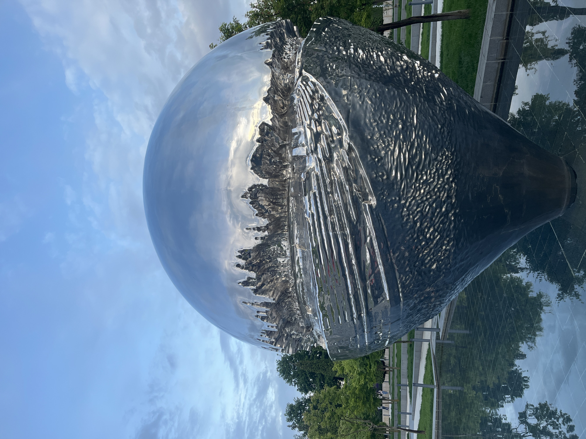 A polished metal sculpture, consisting of a sphere and a conical base, stands near a park. The sculpture's mirrored surface reflects the surrounding environment, including the sky with clouds, trees, and buildings. The sculpture appears to be partially submerged in a shallow pool or reflective surface, which mirrors the trees and the sculpture itself. A metal fence runs along the background, and some greenery is visible. The scene is located in the city of Krasnodar, Russia.