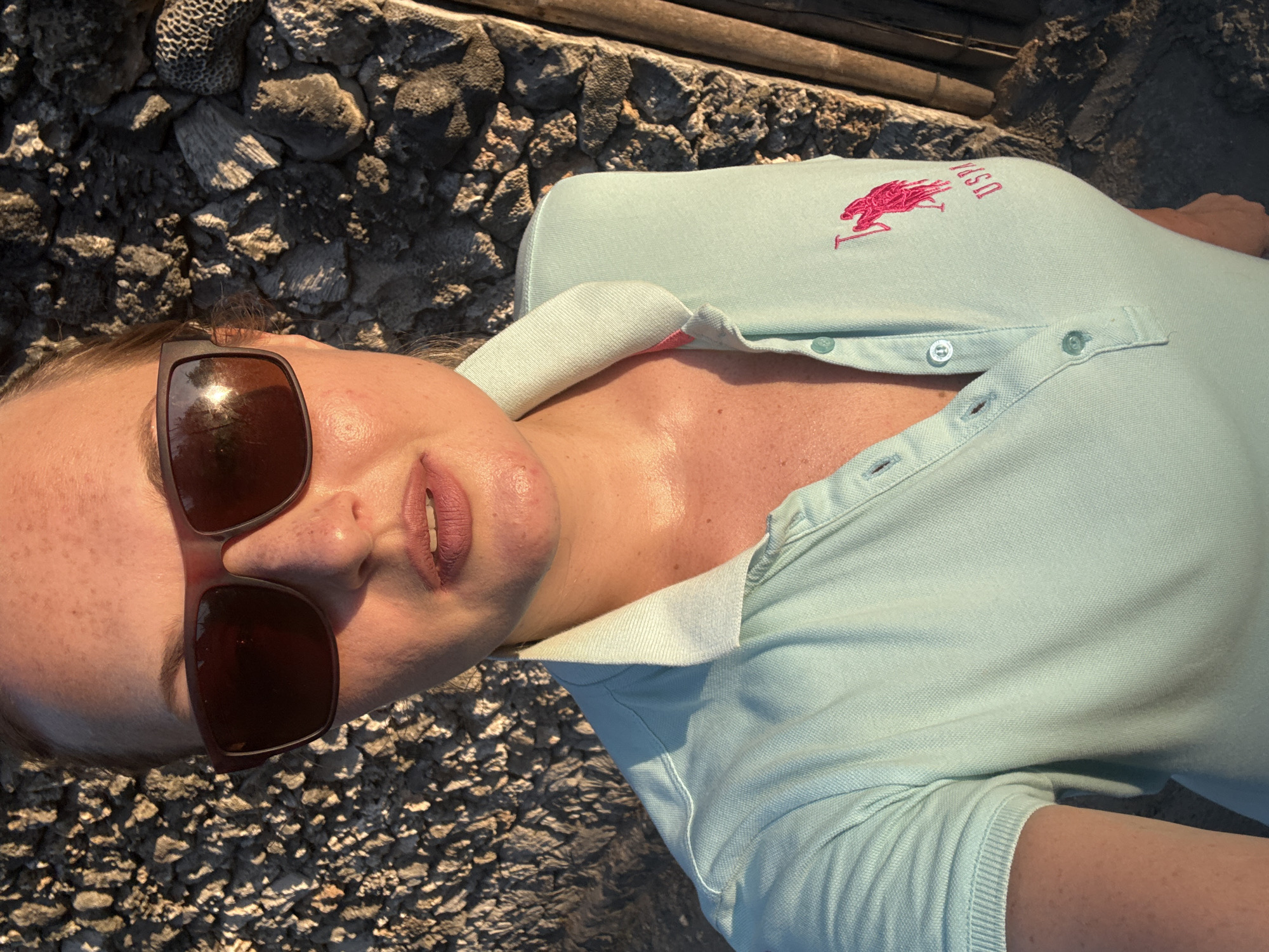 A close-up, selfie-style shot shows a fair-skinned woman with freckles and light brown hair pulled back, wearing oversized sunglasses with dark lenses. She is wearing a light blue polo shirt with a pink logo and lettering on the right chest. The background consists of a rough, textured stone wall and some dried, dark wooden elements. The lighting suggests it is late afternoon or early evening, casting warm, golden light on the woman's face. The overall impression is a casual, outdoor portrait. The specific location in Denpasar, Indonesia, cannot be determined from the visual information provided.