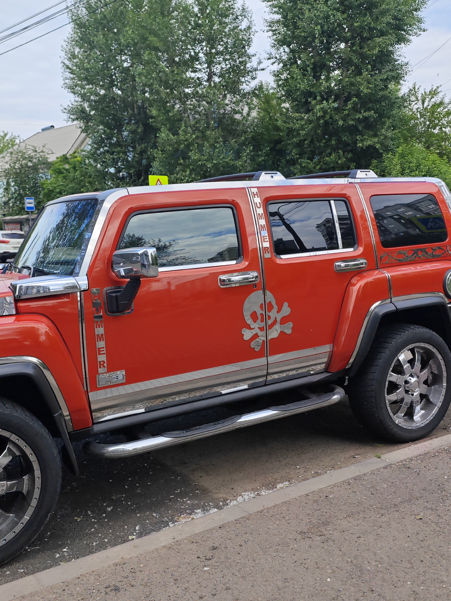 An orange Hummer H3 SUV is parked on a street in Krasnoyarsk, Russia. The vehicle has chrome trim and a skull decal on the passenger side door. The street is paved with asphalt and bordered by a concrete curb. Trees and buildings are visible in the background. The sky is cloudy.