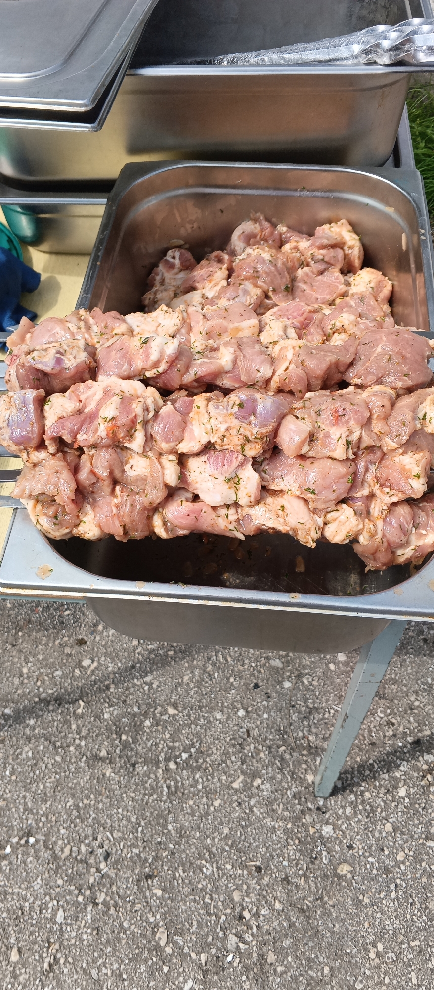 A close-up, eye-level shot shows several metal skewers loaded with marinated raw meat, arranged in a stainless steel food pan. The meat appears to be pork, cut into bite-sized pieces and seasoned with herbs and spices. The food pan is resting on a metal stand, which is partially visible at the bottom of the frame. The background consists of a concrete surface and some green grass. The lighting suggests it is daytime. The scene depicts food preparation, likely for a barbecue or outdoor event, in an unspecified urban location.