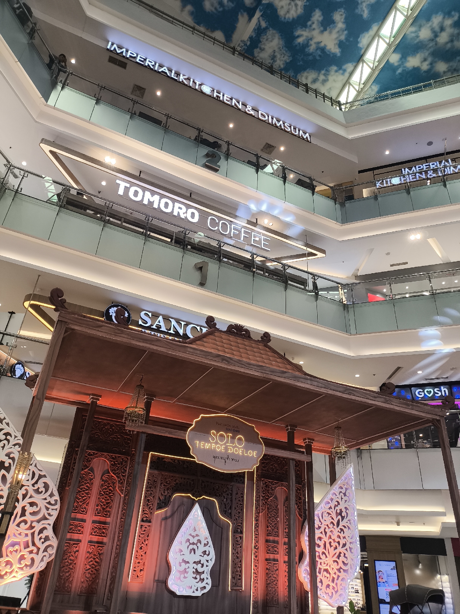 The image depicts an interior view of a multi-level shopping mall, likely in Grogol, Indonesia, given the context provided by the "SOLO TEMPOE DOELOE" signage. The perspective is from a lower level looking upwards, revealing multiple floors with glass railings and neon signs. Prominently displayed on different levels are illuminated signs for "IMPERIAL KITCHEN & DIMSUN" and "TOMORO COFFEE". In the foreground, a decorative wooden structure resembling traditional Indonesian architecture, possibly a booth or display, is visible. This structure is adorned with intricate carvings and features illuminated signage that reads "SOLO TEMPOE DOELOE" and some script in Javanese. Ornate lamps hang from the ceiling of this structure. White, patterned decorative elements, resembling stylized wings or batik motifs, are placed on either side of the wooden structure. The ceiling of the mall is designed to resemble a sky with clouds, illuminated by natural light filtering through a skylight structure at the top. The overall atmosphere suggests a modern shopping environment with a touch of cultural heritage incorporated into its design. No individuals are clearly discernible in the image.