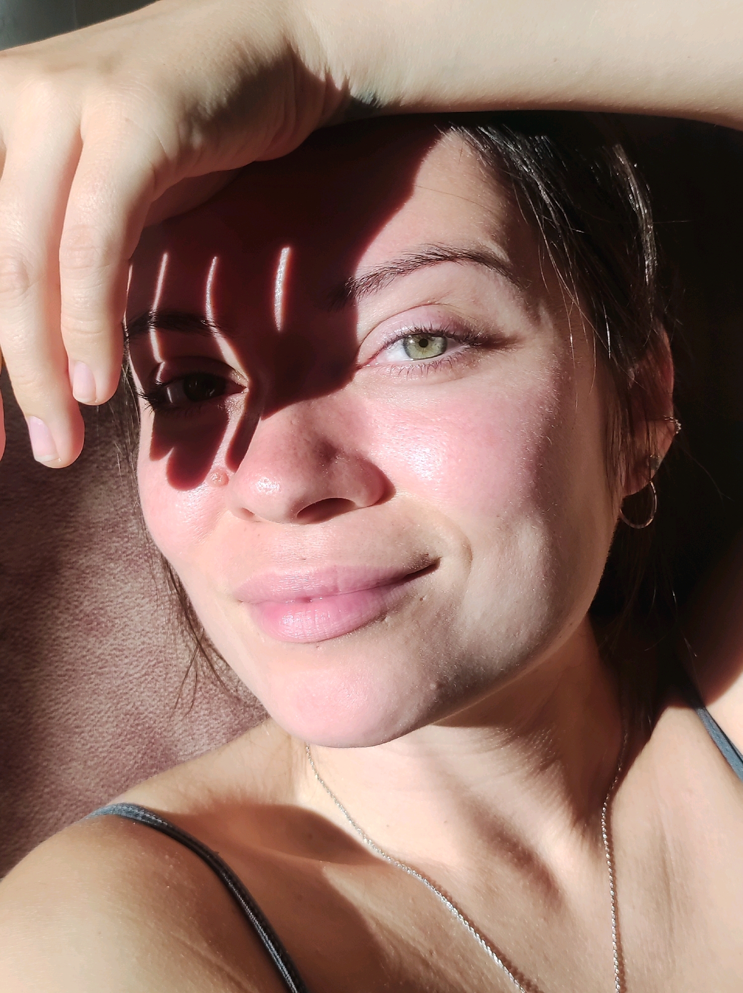 A young woman with light skin and green eyes is lying down, looking up at the camera. Her right hand is raised and resting on her forehead, casting a shadow across her face. Sunlight streams in from the left, creating strong highlights and shadows. She has a faint smile on her lips, and she is wearing a thin necklace. The background is a textured surface, possibly a blanket or couch. The image appears to be a selfie taken indoors.