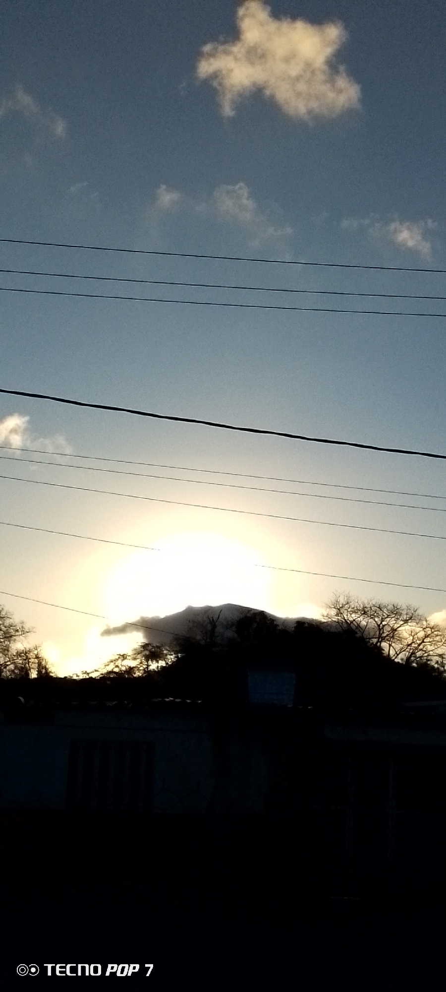 The image shows a sunset scene in Altagracia, Venezuela. The sun is setting behind a dark silhouette of a hill with trees, creating a bright glare. Several utility lines run across the sky. Above the lines, the sky is pale blue with a few scattered clouds. In the foreground, the silhouette of a building is visible. The "Tecno Pop 7" watermark is in the lower left corner.
