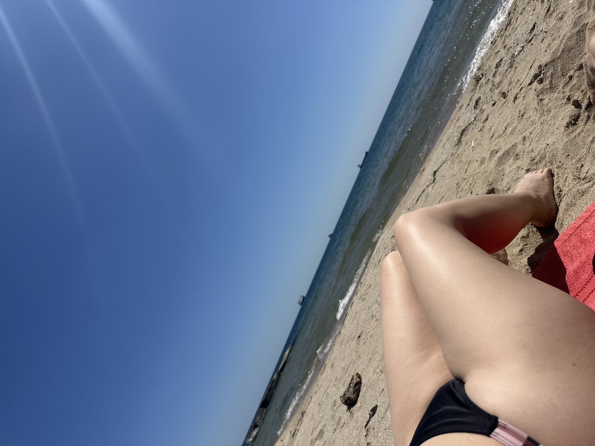 Image depicts a sunny day at the beach, likely in Varna, Bulgaria, considering the submission context. Clear blue sky dominates the left side of the image with visible sun glare. A sandy beach leads to the sea, visible in the upper center of the frame. The water is calm with small waves breaking on the shore. A dark structure, possibly a breakwater or old pier remains, is visible in the water along the shoreline. In the lower right of the image, a person is reclining on the beach on a red towel, wearing a black bikini bottom with pink accents. Parts of the person's legs and feet are visible. The sand is textured with footprints and occasional debris like a small piece of wood.