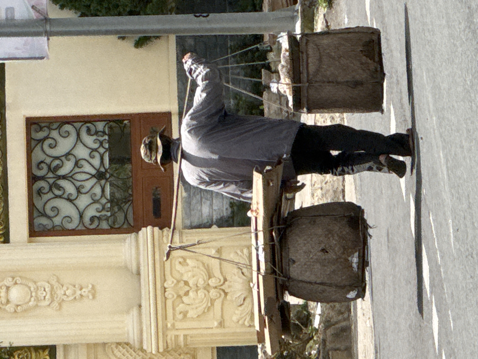 A man wearing a wide-brimmed hat and a grey long-sleeved shirt walks from left to right on a paved road, carrying a yoke across his shoulders. Suspended from the yoke by ropes are two large, woven baskets filled with what appear to be goods or produce. The baskets are dark brown and weathered. The man is wearing dark trousers and sandals. The background features a building with ornate architectural details, including columns and decorative carvings, and a window with a wrought-iron grille. A light pole is visible on the right side of the frame. The scene appears to be in Da Lat, Vietnam, characterized by the colonial-influenced architecture and the presence of a street vendor.
