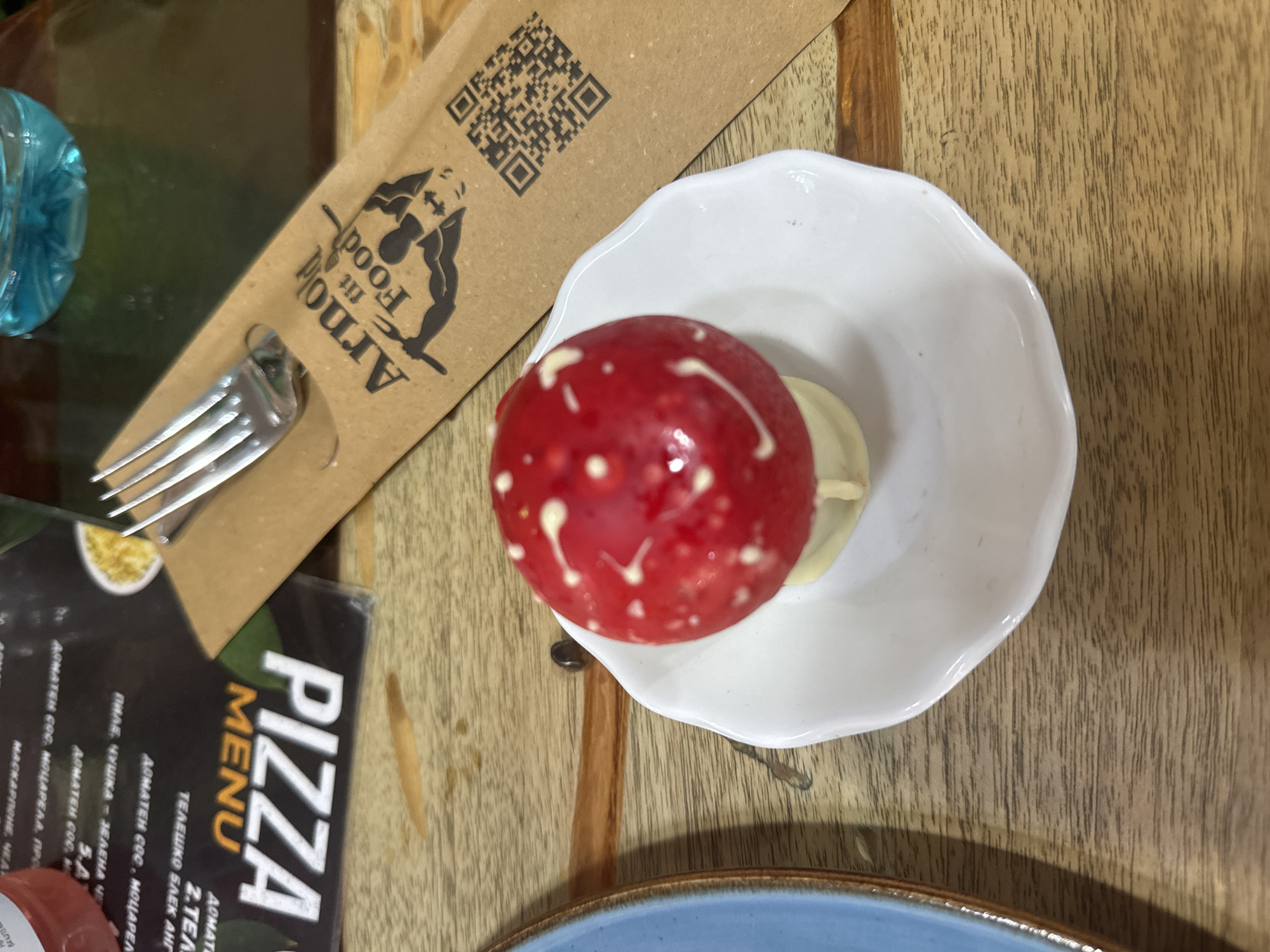 The image shows a still life on a wooden table. A red, spherical dessert with white dots sits on a small, decorative white plate. To the left is a paper utensil holder with the logo of "Arnold fit Food" and a QR code. Next to the holder, there is a fork and a corner of a "Pizza Menu" in Bulgarian. A blue water bottle is partially visible on the left. A blue-rimmed plate is partially visible at the bottom. The photo appears to have been taken in Sofia, Bulgaria, based on the Bulgarian text on the pizza menu.