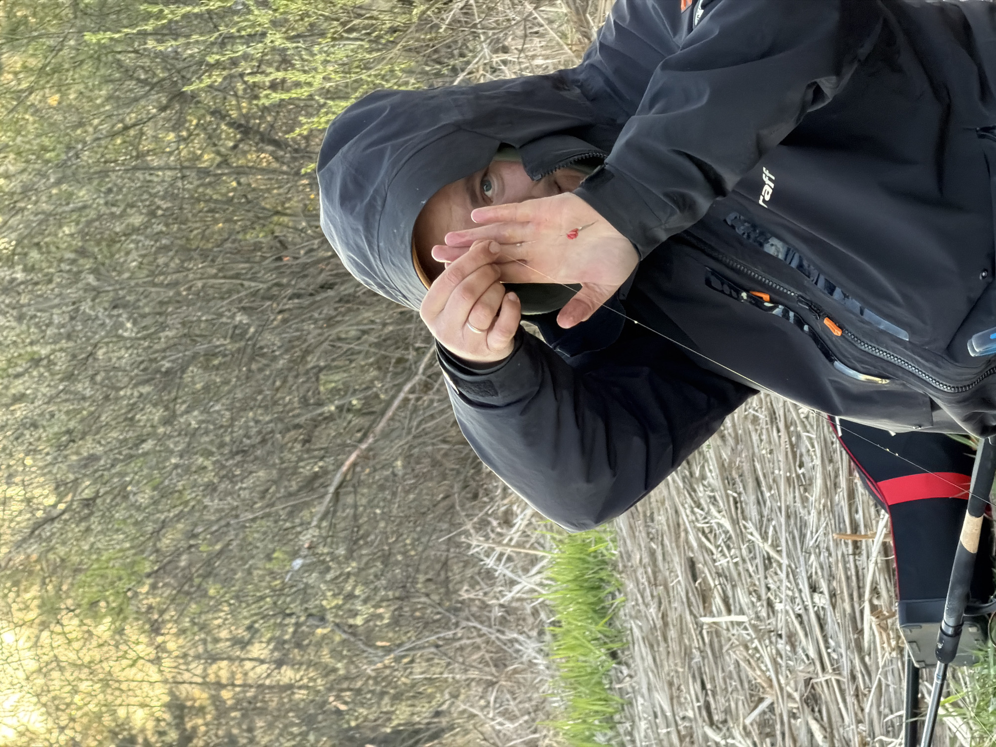 A person wearing a black hooded jacket is holding a fishing line with a red bobber or float attached. They are outdoors, possibly near a body of water, as suggested by the tall grass and reeds in the background. The location is likely Chaikovsky, Russia, based on the prompt.