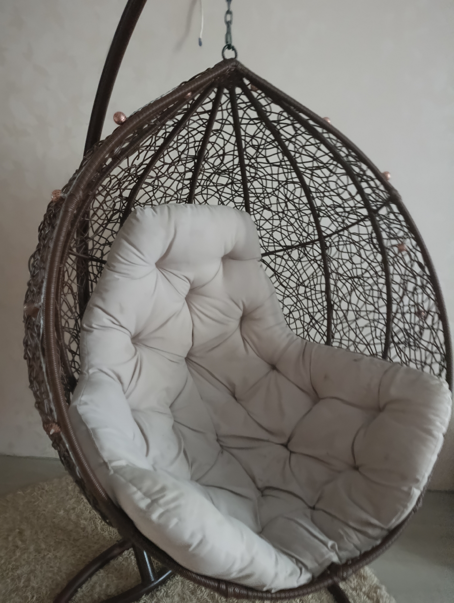 The image shows a brown wicker hanging egg chair with a light beige cushioned seat, indoors. The chair is suspended from a dark brown metal frame, which is supported by a stand with two legs visible on a textured beige rug. The background is a plain, light-colored wall. There are no people in the image. The scene is an interior shot and does not provide any information about the city of Kharkiv, Ukraine.