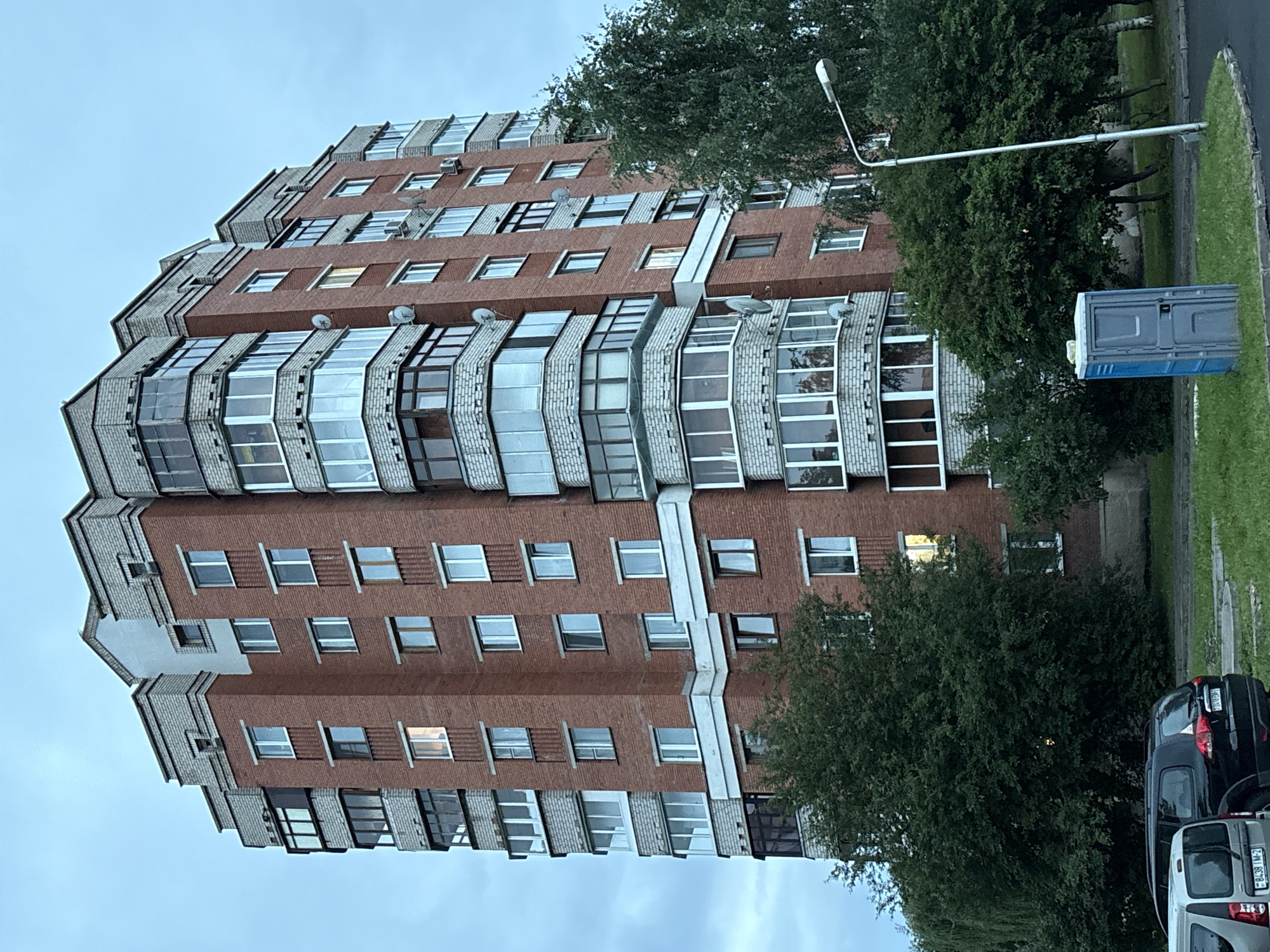 The image depicts a multi-story red brick apartment building with white decorative elements and numerous windows, some of which are bay windows with glass enclosures. The building is surrounded by green trees and a grassy area. In the foreground, a portable blue toilet is visible on the grass, next to a streetlight. Several cars are parked in the lower left portion of the image. The sky is overcast and a pale blue-gray. The overall scene appears to be an outdoor urban environment.