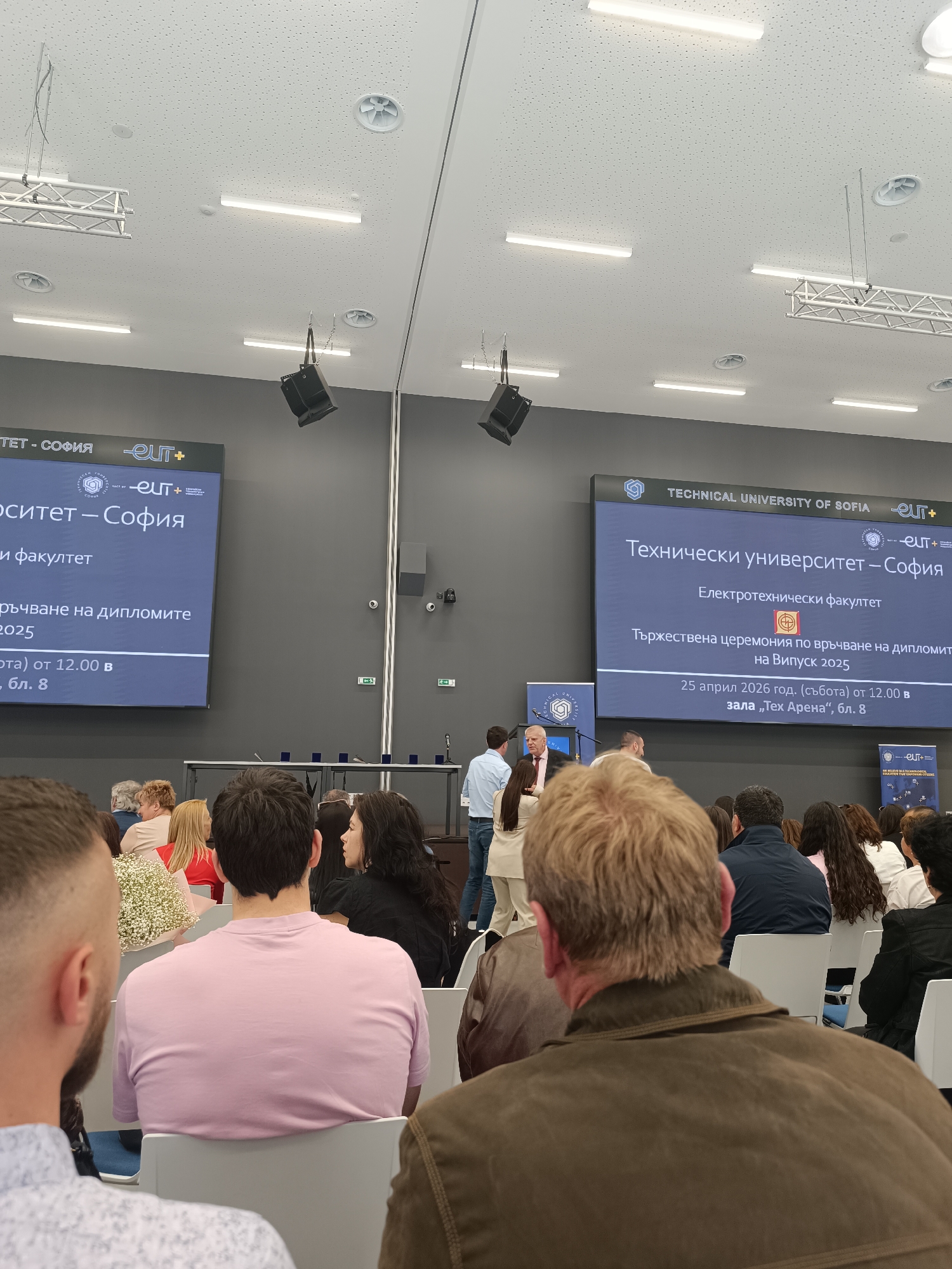 This image shows an audience seated at a ceremony held at the Technical University of Sofia in Sofia, Bulgaria. On a large screen in front of the audience, the information indicates a ceremony for the graduation class of 2025, scheduled for April 25, 2026, at 12:00 in "Tech Arena," block 8. Several individuals are visible in the foreground and background, seated in rows of chairs facing the stage where the screen is located. Speakers are visible at the stage. The hall has a high ceiling with lighting fixtures and speakers. The room appears to be well-lit, and the overall setting suggests a formal event or gathering.