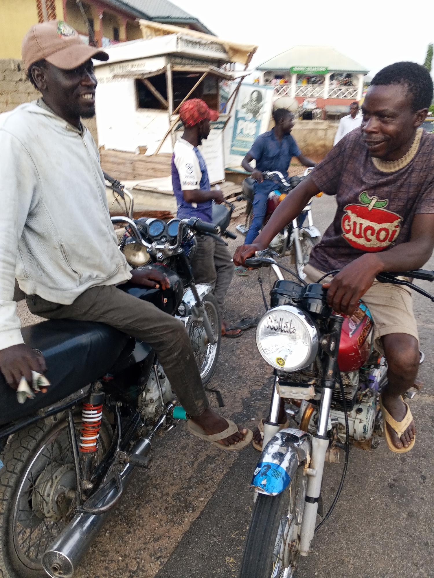 The image captures a vibrant street scene in Zango-ndaji, Nigeria, featuring several men gathered with their motorcycles under soft daylight. The atmosphere appears friendly and relaxed, with the main subjects smiling.
In the immediate foreground on the left, a man with dark skin, wearing a light-colored hooded sweatshirt, dark trousers, and a brown baseball cap, sits astride a black motorcycle. He faces right, smiling broadly, with his left hand resting on the seat, holding some crumpled material. His right hand is on the handlebars.
To his right, another man with dark skin, also smiling, is seated on a red and chrome motorcycle. He wears a dark brown t-shirt featuring a prominent red apple graphic with the word "Gucci" written across it, along with light-colored shorts and flip-flops. His hands are on the handlebars. Notably, his motorcycle's chrome headlight bears Arabic script, which reads "ما شاء الله" (Masha'Allah). A blue sticker with white text and a face, possibly related to a public health campaign like COVID-19, is affixed to the front fender.
In the background, several other men are visible. One man in a blue and white t-shirt and a red cap is on another motorcycle behind the left foreground subject. Further back, another man in a blue shirt is on a motorcycle near a sign that clearly displays "POS." A building with a green roof and a white railing balcony, along with a small white kiosk-like structure, completes the backdrop. The scene suggests a casual gathering or a brief stop along a paved road in a local community, typical of areas where motorcycles are a primary mode of transport. The soft lighting indicates either early morning or late afternoon.