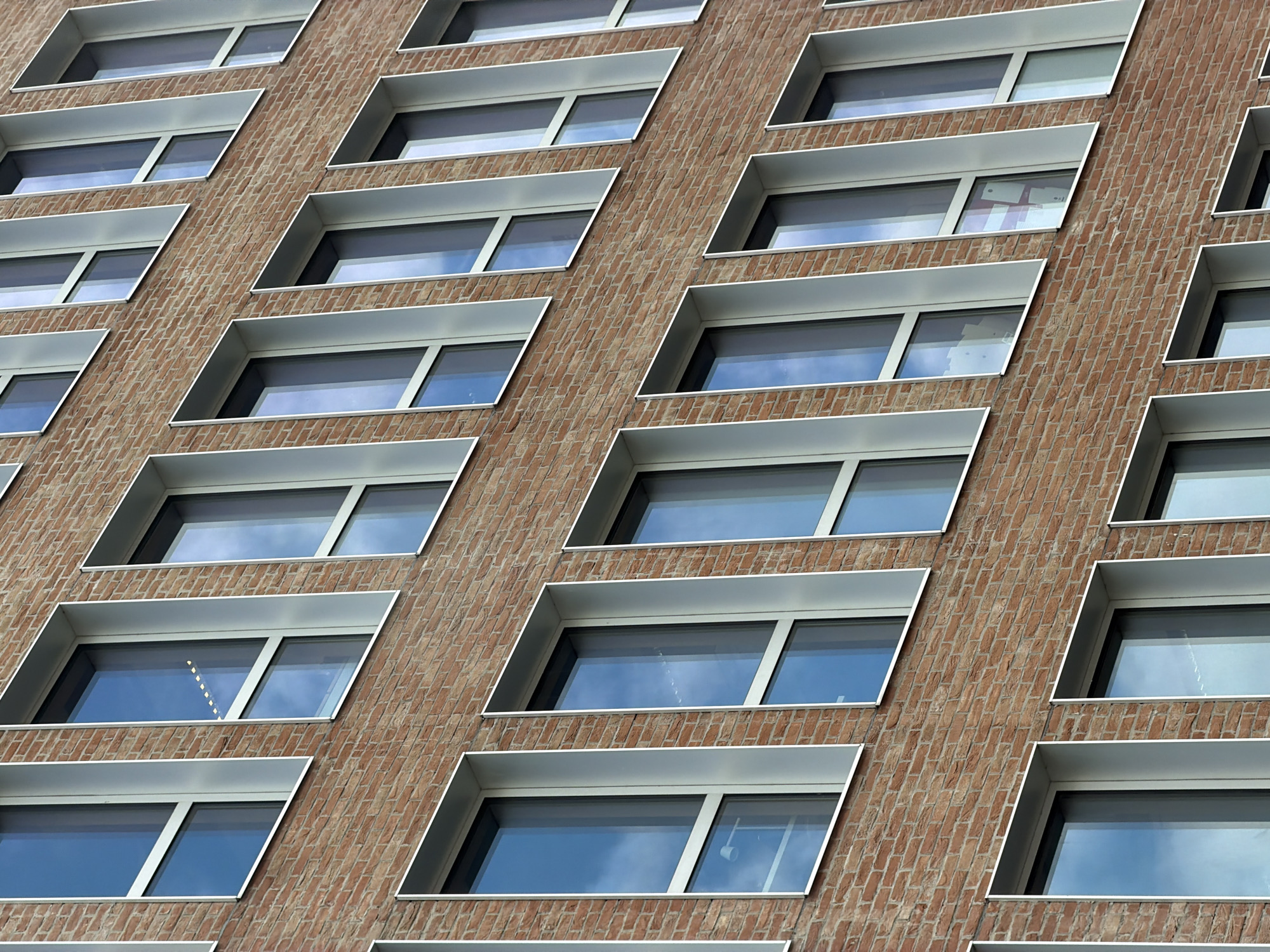 The image shows a close-up, angled view of a modern building facade in Ivry-sur-Seine, France. The facade is constructed from reddish-brown brick with a repeating pattern of rectangular windows. Each window is framed by a light-colored, angled metal trim, creating a distinctive geometric design. The windows themselves are reflective, showing a bright, overcast sky with hints of blue. Some of the windows appear to be offices or living spaces, with glimpses of interior lighting and subtle reflections of what might be papers or screens. There is no visible text in the image. The scene is likely captured during daylight hours, given the brightness of the sky reflected in the windows. The architectural style suggests a contemporary urban environment.