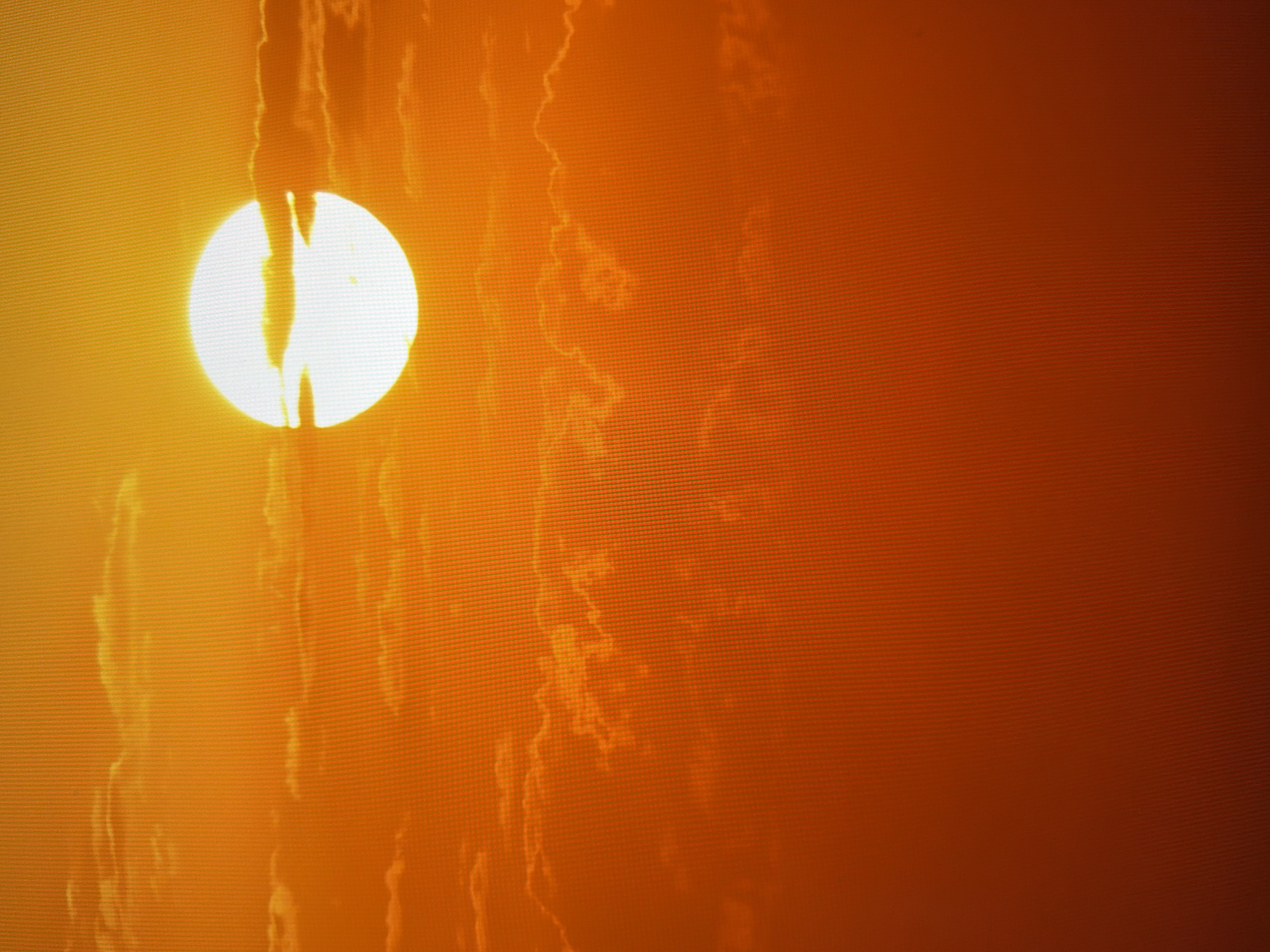 The image shows a bright, full sun partially obscured by thin, wispy clouds during what appears to be sunset or sunrise. The sky is a vibrant gradient of orange and yellow, with some deeper red tones in the lower part of the frame. The sun itself is a brilliant white disc with a distinctly illuminated edge. The clouds are tinged with the colors of the sky, creating a dramatic and warm atmosphere. The surface of the image exhibits a subtle, fine grid pattern, characteristic of a screen display. There are no people, animals, or man-made structures visible in the image. The scene is natural and atmospheric, with a focus on the celestial event. There is no information within the image that allows for the identification of a specific location in Kyiv, Ukraine.