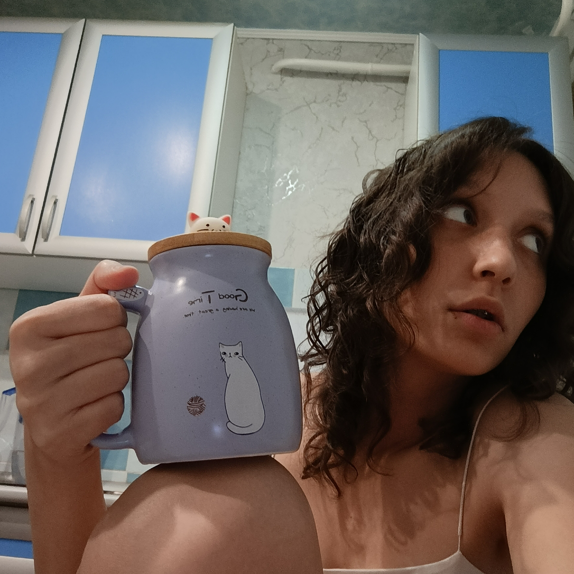 A young woman with curly brown hair is holding a light purple mug in her left hand. The mug is resting on what appears to be her shoulder or upper chest. The mug has a wooden lid with a small white cat figurine on top. The mug also features a white cat drawing and the words "Good Time" written on it. The woman is looking to her right with a slightly surprised expression. She is wearing a white tank top. The background includes what looks like kitchen cabinets with blue sky visible through the glass panels and a wall with a marble-like pattern. The location is inside a building, possibly a kitchen.