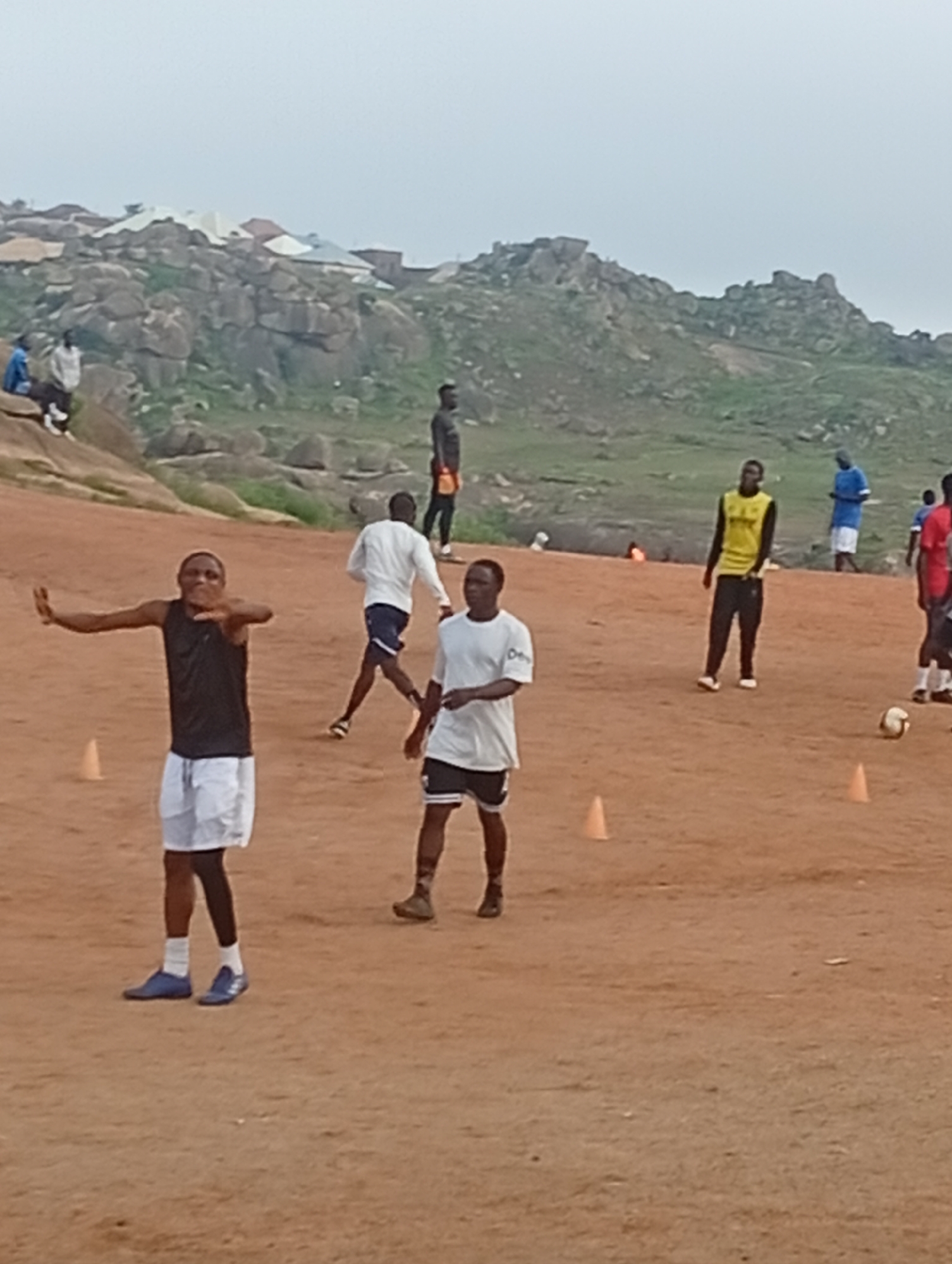The image captures a group of men engaged in what appears to be a soccer practice or game on a dusty, reddish-brown field. The terrain is uneven and interspersed with small orange cones, suggesting a training drill. In the background, a rugged, rocky hillside with scattered buildings and houses is visible, characteristic of parts of Jos, Nigeria. The sky is overcast, creating a diffused, soft light.
In the foreground, a man in a black sleeveless shirt and white shorts is positioned centrally, his arms outstretched and hands open, as if in mid-action or giving instruction. To his right, another man in a white t-shirt and dark shorts is running. Further back, a man in a yellow vest and black pants stands still, facing away from the camera. To his left, a man in a blue shirt and white shorts walks away. Other figures are scattered across the field, some further in the distance, wearing various athletic attire, including red and blue shirts. A soccer ball is visible on the ground near the man in the yellow vest. The overall scene depicts a casual outdoor sports activity in a natural, somewhat undeveloped environment.