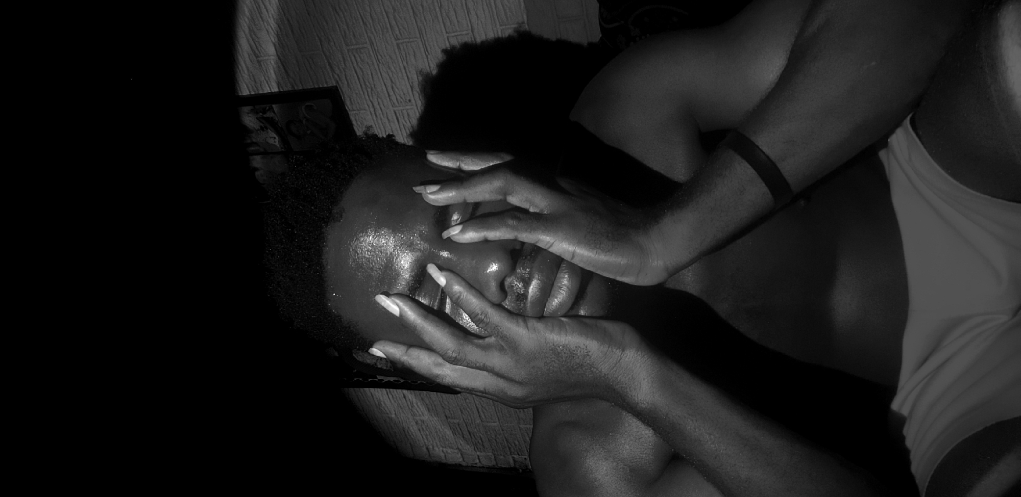 A black and white photograph, taken in Narayi, Nigeria, captures a person with dark skin lying down in a dimly lit indoor setting. The person's head is positioned towards the left of the frame, with their body extending to the right. Both hands are raised and cover the majority of their face, from the forehead down to the mouth, obscuring their eyes and nose. This gesture conveys emotions such as distress, contemplation, exhaustion, or a desire for privacy.
The person's skin appears lustrous or wet, catching the dramatic low-key lighting and creating highlights that emphasize the contours of their face and hands. They have natural, dark, curly hair. A dark band is visible on their left arm. The upper body appears bare, while a light-colored garment, possibly shorts or underwear, covers the lower torso on the right side of the frame.
The background includes a textured surface, possibly a patterned wall or fabric, visible behind their head and upper body. A small, framed object can be faintly seen in the upper left background, near the person's head. The rest of the background, particularly on the far left, is dominated by deep, dark shadows, contributing to the intense and intimate atmosphere. The time of day is suggested to be night or a very dim interior due to the lighting. There is no visible text in the image.