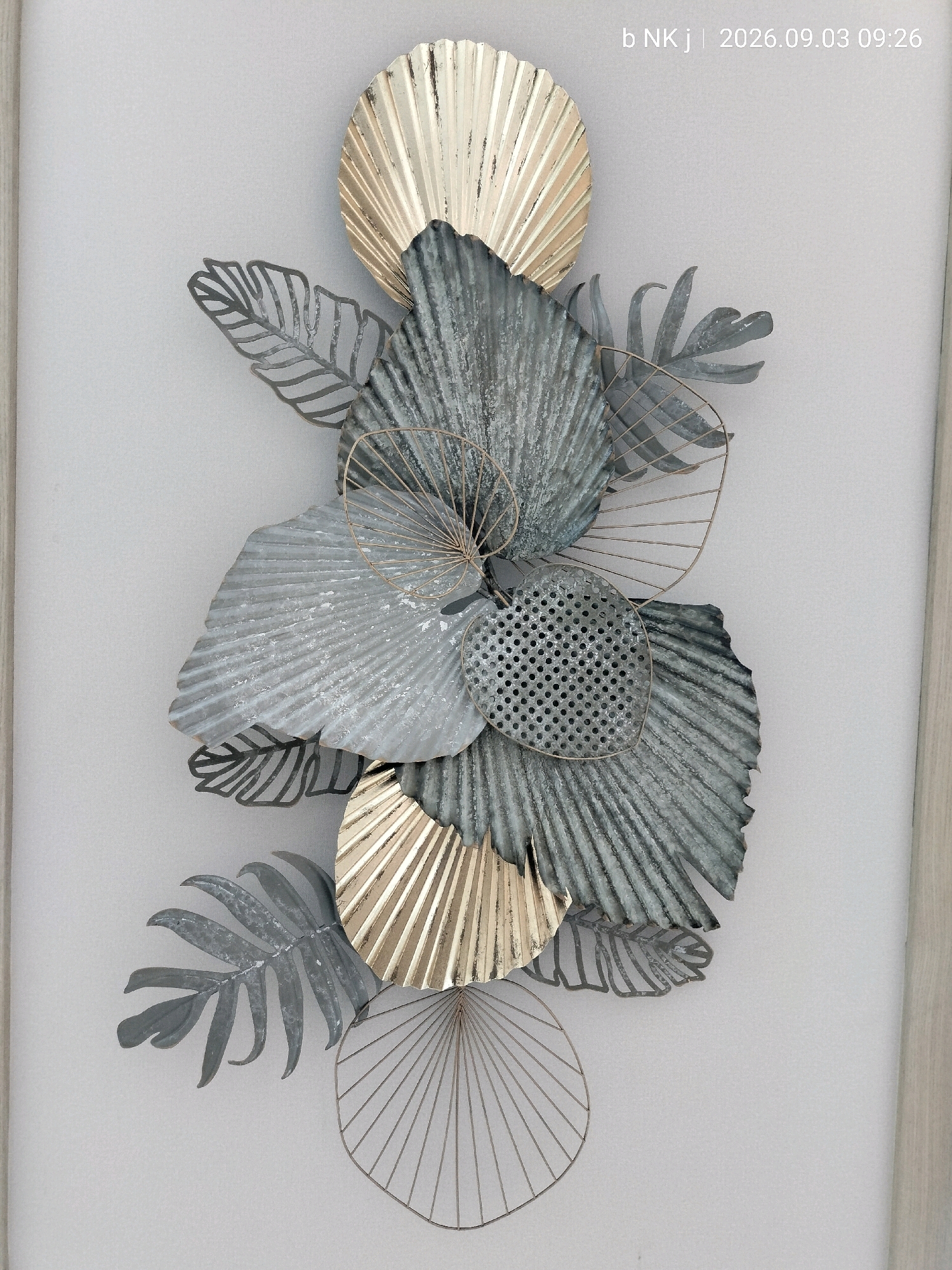 A detailed, full-frame, eye-level shot captures a piece of metal wall art displayed against a plain, light-colored wall, likely in South Jakarta, Indonesia. The artwork itself is a multi-layered arrangement of stylized botanical elements. Dominating the composition are several fan-shaped elements, some appearing to be palm fronds, crafted from corrugated metal with a distressed, weathered finish in shades of grey and metallic silver. Interspersed with these are elements resembling fern leaves, also made of metal, some with a solid silhouette and others rendered as delicate, etched outlines. Two prominent fan shapes at the top and bottom of the arrangement are finished in a metallic gold, adding a contrasting warmth to the cooler grey tones. One of the grey fan elements in the center features a perforated surface, creating a subtle visual texture. The artwork is characterized by its metallic sheen and the interplay of solid and openwork forms. The background wall is uniformly lit, with a subtle shadow cast by the wall art, suggesting it is mounted at a slight distance from the surface. A narrow vertical element, possibly a doorframe or molding, is visible on the far right edge of the image, adding a subtle vertical line to the composition. The lighting is diffused, with no harsh shadows, indicating an indoor environment. The overall impression is one of elegant, contemporary decor.