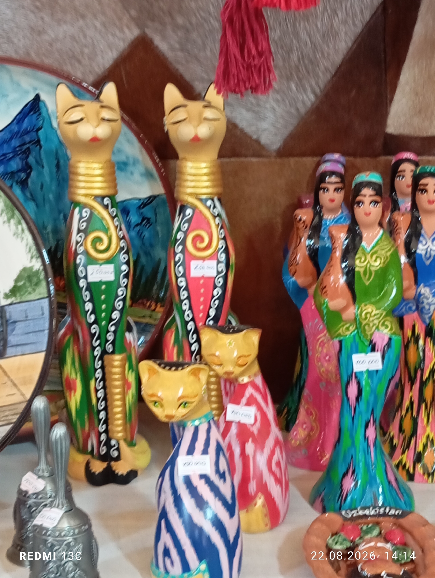 The image displays a collection of decorative items, likely souvenirs, arranged on a shelf or table. In the foreground, to the left, are three silver-toned bells, two of which are clearly visible. The closest bell has a label with the text "REDMI 13€". To the right of the bells, and slightly further back, are two ceramic cat figurines. The left cat is predominantly green with black, white, and red decorative swirls, and a yellow base. It has a small white label attached to its body. The right cat is primarily red with white and black decorative patterns, also with a yellow base and a similar white label.
Behind these initial items, a group of ceramic doll figurines dressed in traditional Uzbek attire are visible. These dolls are characterized by their colorful, patterned dresses and headscarves. One doll on the far right wears a vibrant, multicolored patterned dress. A doll in the center wears a bright green dress with intricate gold embroidery and a blue and turquoise headscarf, holding a brown pot. Another doll to its left wears a pink dress.
Further back and to the left, two taller, slender cat figurines stand out. The left cat is green with detailed black and white swirling patterns and yellow accents, adorned with gold bands around its neck and midsection. It also has a white label. The right cat features a predominantly red body with similar black and white decorative patterns and gold neck adornments.
The background is a textured surface, appearing to be a tapestry or rug with shades of brown and grey, and a hint of red fringe hanging from the top. A partially visible decorative plate or mirror with a blue landscape design is situated behind the left green cat.
The timestamp "22.08.2026, 14:14" is visible in the bottom right corner, suggesting the photograph was taken on this date and time. While the image is described as being from Amirsay, Uzbekistan, this specific location information is not directly evident from the visual elements within the photograph itself, other than the style of the dolls and decorations which are indicative of Uzbek craftsmanship.