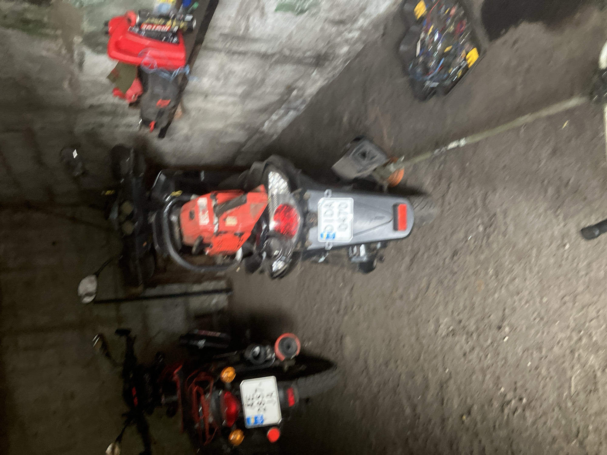 This is a forensic, matter-of-fact description of an indoor scene, likely a garage or shed, located in Vovkivka, Ukraine. The scene contains two motorbikes and various tools.
In the foreground, a dark-colored motorbike is parked. It has a license plate with the characters "KE 2857 JA". A red chainsaw is mounted on the rear of this motorbike. The motorbike also features orange turn signals and a red taillight.
To the right of the first motorbike, a second, lighter-colored motorbike is visible. Its license plate displays "DIDA 0470". This motorbike also has a red taillight.
In the background, a shelf is mounted on a rough, textured wall. On the shelf, there are several items, including a red fuel canister and several spray cans.
On the floor to the right of the motorbikes, there is a collection of tools, including what appears to be a grass trimmer with a long shaft extending upwards and to the left, and a black container filled with various items, possibly electrical components or smaller tools.
The environment is dimly lit, with a rough, dirt or concrete floor. The walls appear to be made of concrete or a similar rough material, with some areas showing a greenish tint. The overall impression is that of a storage area for vehicles and tools.