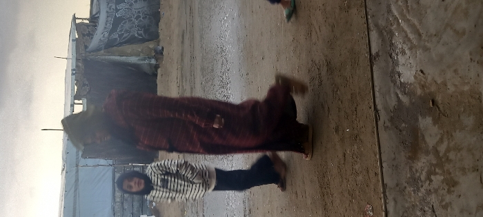 The image depicts an outdoor scene in Khan Yunis, Palestinian Territory, under an overcast sky. The ground is an uneven mixture of damp earth and mud, with a wet, dark, possibly hardened or paved strip visible across the lower foreground. In the background, two makeshift structures stand: one on the left appears to be a shelter constructed from light-colored tarpaulin stretched over a frame, and another on the right is a darker, weathered tent-like dwelling made of patched materials, possibly with a decorative dark patterned fabric draped over part of it. Visible support poles are present on both structures.
Three individuals are in motion, walking from right to left across the scene. A young individual, likely a child or adolescent, is positioned towards the left foreground. They are wearing a black and white horizontally striped hooded top, dark trousers, and brown shoes, with their hands in their pockets and their gaze directed forward. To their right and slightly behind, an adult figure is largely blurred due to motion, wearing a long, dark red patterned coat or dress, a head covering, and light-colored shoes. On the far right, only the lower leg and foot of a third individual are visible, wearing a light green sandal.