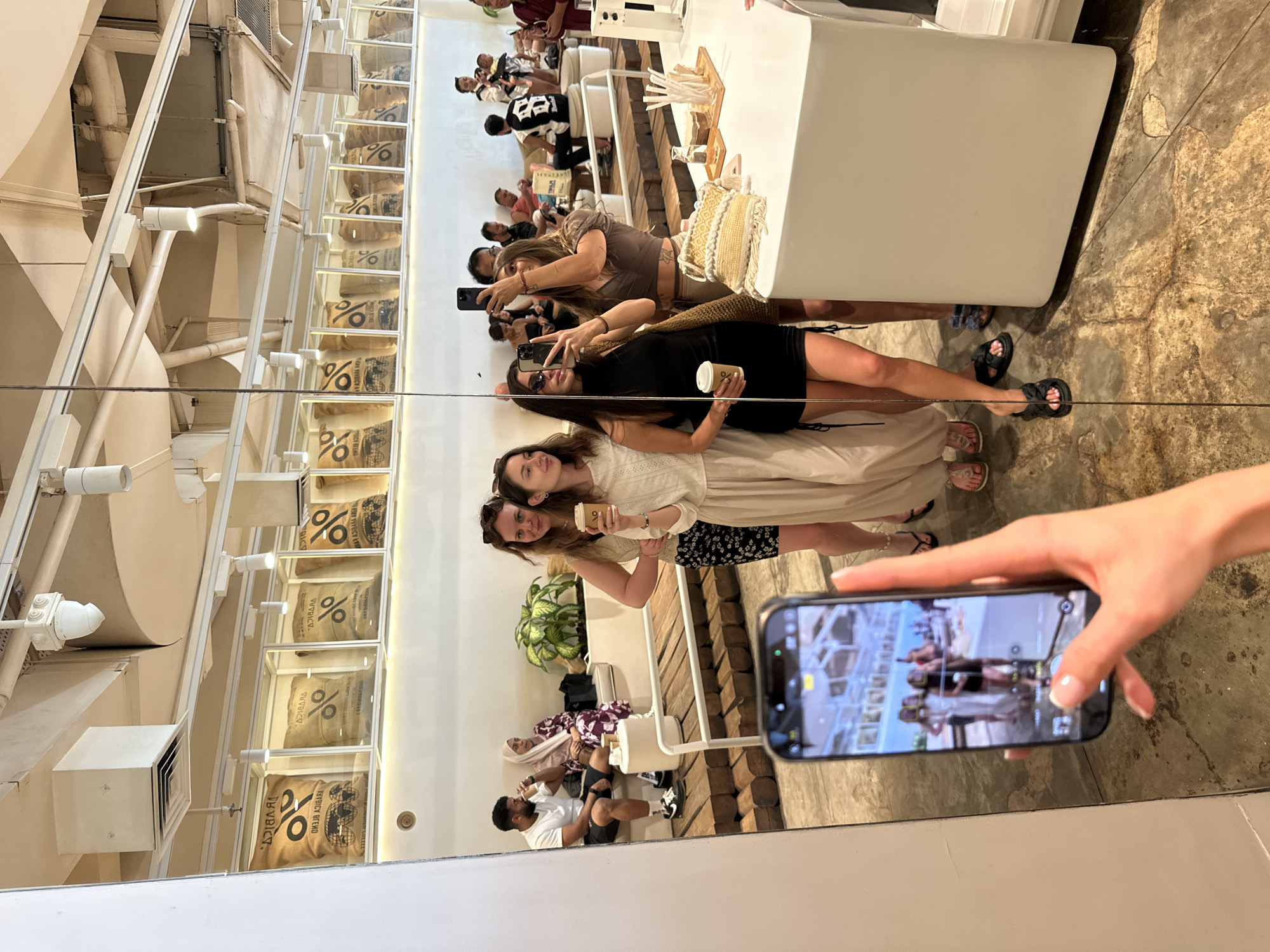 Here's a detailed description of the image:
**Setting:** The photo appears to be taken in a mirror, likely inside a % Arabica coffee shop. This conclusion is supported by the presence of coffee beans displayed in glass containers with the '%' symbol and a counter typical of a coffee shop.
**People:** Several people are present. Two women are standing close together, with one holding a coffee cup. There's another woman behind them, holding her phone up, likely taking a picture. Additional people are visible in the background and in the reflection of the hand holding the camera of the mirror.
**Objects:**
* A white counter is visible, stocked with coffee shop supplies.
* The coffee bags are displayed, with the distinctive '%' logo.
* A hand is holding a phone, taking a picture of the scene in the mirror.
**Actions:** People are taking photos and posing. The hand is capturing the scene in the mirror.
**Interactions:** People are interacting by taking pictures of each other and posing.
