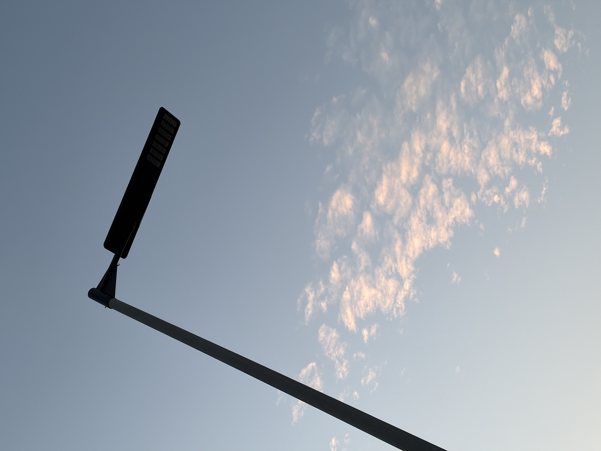 A street lamp is silhouetted against a clear blue sky in Igboefon, Nigeria. The lamp post is dark gray and extends from the bottom left corner of the frame diagonally upwards. At the top of the post, the lamp itself is a long, rectangular black fixture with multiple small, square lights visible. A thin, scattered band of pale pink and white clouds stretches horizontally across the middle of the frame. The sky is a gradient of light blue. There are no people or animals in the image.