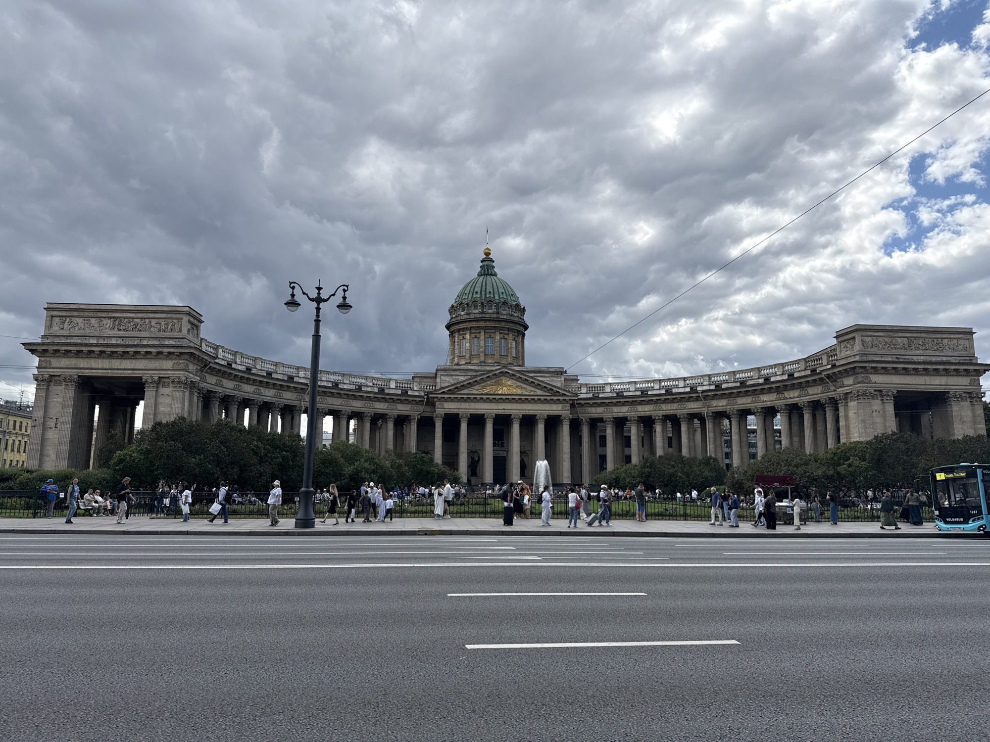 The image depicts a wide, gray sky filled with dramatic, heavy clouds, indicating overcast weather. In the center, a grand, neoclassical building with a large green dome and a colonnade of tall, stone pillars dominates the middle ground. The building has a curved facade, typical of St. Petersburg's imperial architecture. A prominent street lamp with multiple lights stands to the left of the building. In front of the building, a sidewalk area is lined with trees and a metal fence, behind which people are walking and standing. A road with white lane markings occupies the foreground. On the far right, a teal-colored bus with the word "VOLGABUS" is visible, suggesting it is a public transport vehicle. Several people are seen walking on the sidewalk, some appearing to be tourists. The overall scene is that of a public space in the city of St. Petersburg, Russia, likely a popular tourist attraction given the presence of people and the iconic architecture. The building is identifiable as the Kazan Cathedral, a prominent landmark in St. Petersburg.