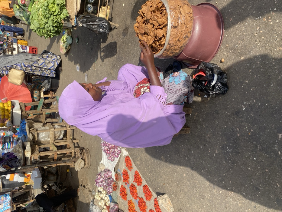 A woman wearing a purple hijab sits among various goods for sale at a marketplace. She sits beside a large container of brown snacks. Other items nearby include produce like lettuce, lemons, onions, garlic, and tomatoes. The market appears to be outdoors on a sunny day, with the shadow of the woman and her goods visible on the pavement.