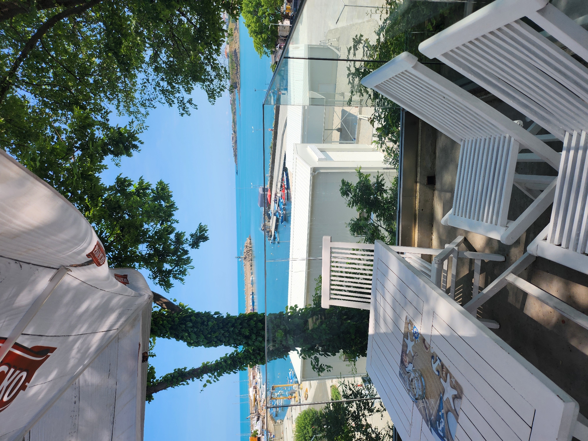 An outdoor seating area with a white wooden table and chairs is positioned on a balcony overlooking a bright blue sea and a harbor. The table is set with a decorative placemat featuring a nautical theme and an ashtray. In the background, several boats are docked in the harbor, and a lighthouse stands at the entrance. The scene is framed by lush green trees and a blue sky. The location is identified as a restaurant in Tsarevo, Bulgaria.