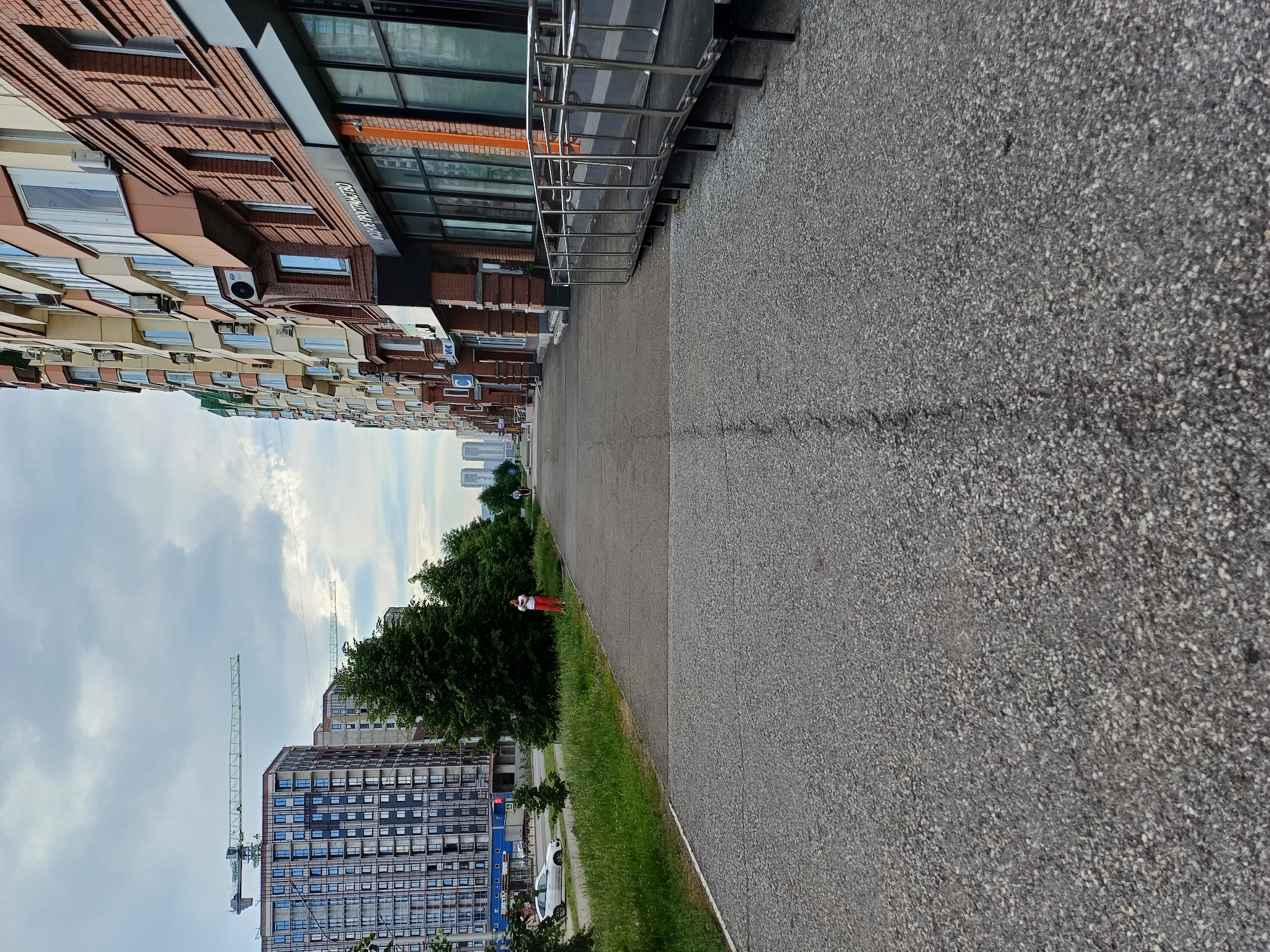 A slightly overcast, daytime exterior shot captures a city street scene. In the foreground, a dark gray asphalt walkway runs diagonally from the bottom left towards the right of the frame. The walkway has a visible seam running down its center and appears slightly worn. To the left of the walkway, a strip of green grass and vegetation lines the edge of the road. Beyond the grass, a white sedan is parked on the left side of the road.
The background on the left features a tall, modern residential building with numerous windows and a construction crane extending from its roof. Another, slightly shorter residential building is visible behind it. Further back, a distant skyline of more tall buildings can be seen under the cloudy sky.
The right side of the frame is dominated by a modern, multi-story building with a facade of brick and cream-colored panels. The building has multiple windows, some of which are covered by air conditioning units. Below the residential floors, there appears to be a commercial storefront with large glass windows. A metallic ramp with a railing extends from the sidewalk towards the street.
In the mid-ground, near the edge of the grass, a person wearing red pants and a white top is standing. Further down the walkway, another person is visible riding what appears to be a scooter or bicycle. Trees are interspersed throughout the scene, with dense green foliage. The overall impression is a developed urban area with ongoing construction and pedestrian activity.