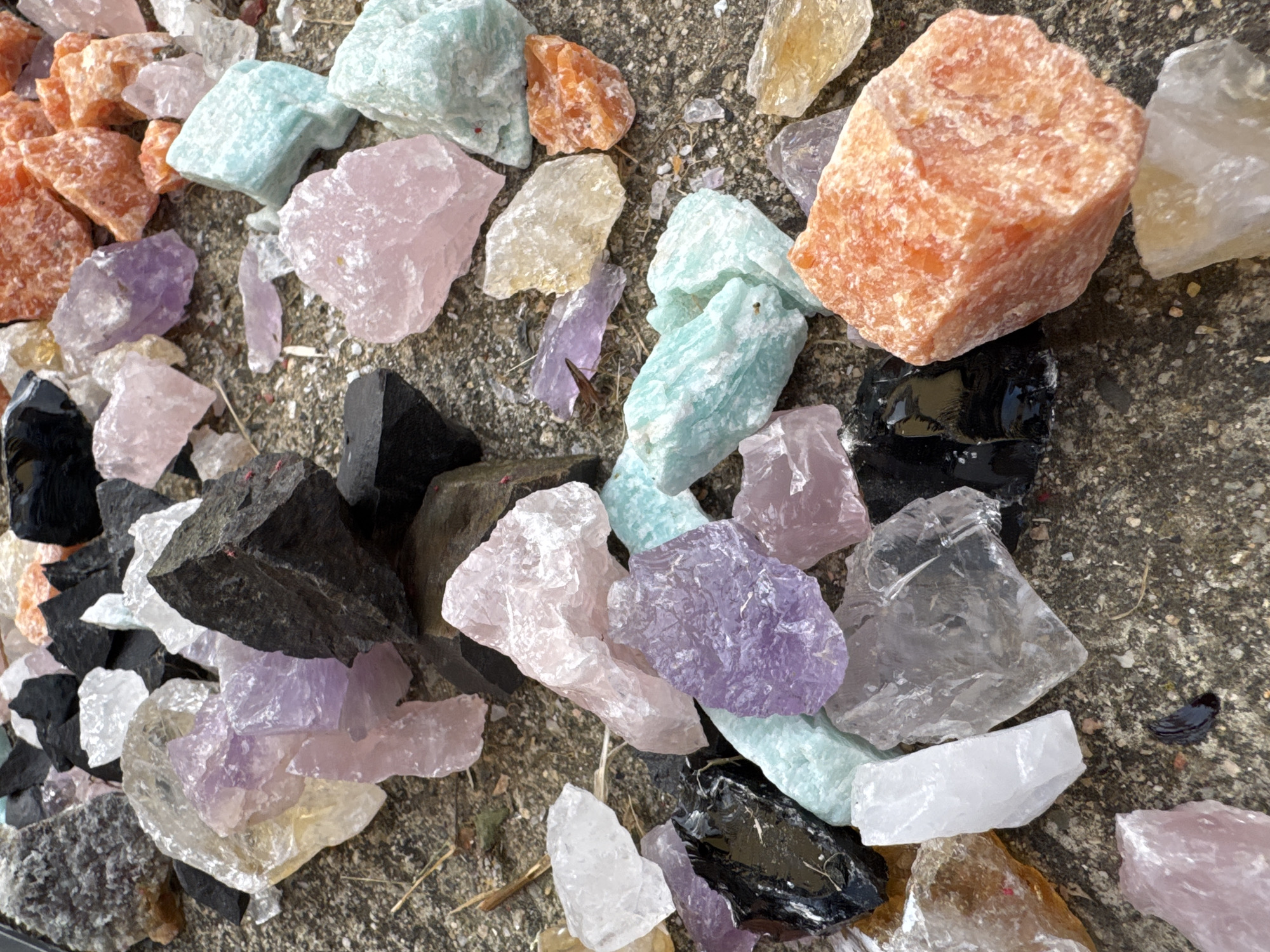 The image shows a collection of rough-cut stones of various colors and sizes scattered on a weathered concrete surface. The stones include shades of orange, blue, pink, purple, clear, and black. The surface shows signs of wear and possibly exposure to the elements. The location is not identifiable based on the image.